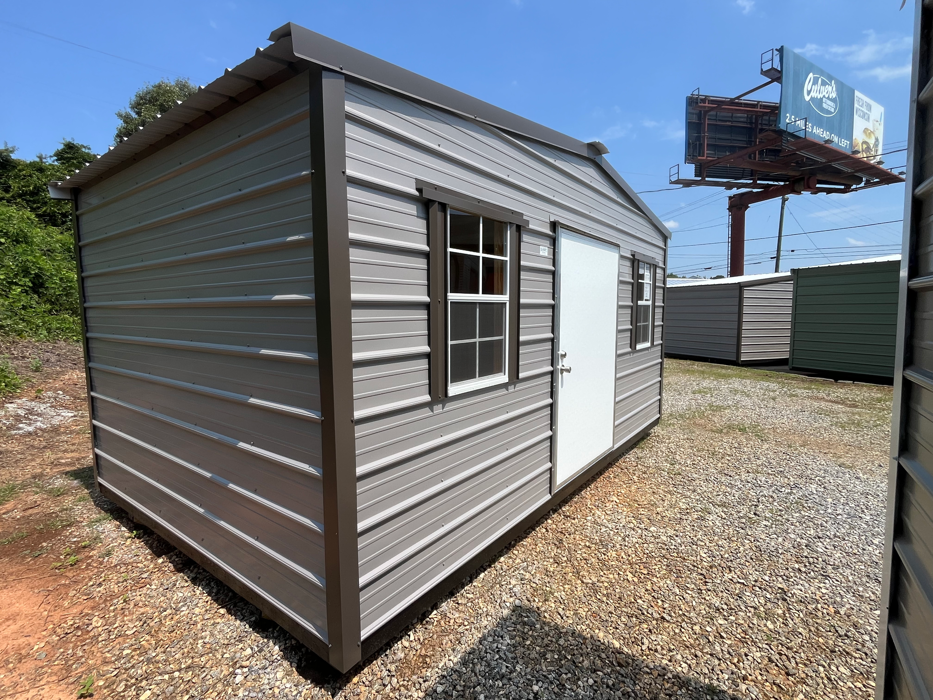 Small gray metal shed with a window and white door on a gravel lot under a blue sky.