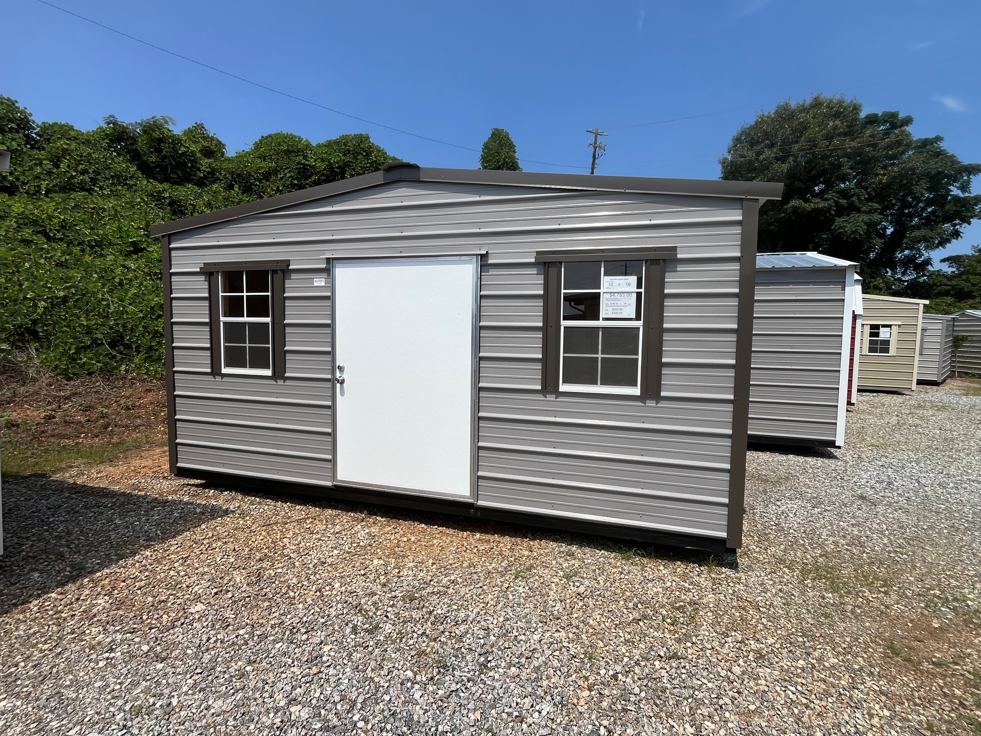 Gray metal storage shed with white door and windows on a gravel lot under a blue sky