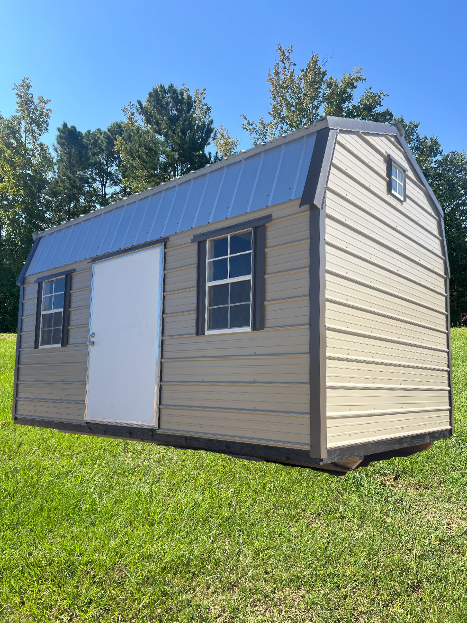 Tan barn-style shed with blue roof and white door and windows on green grass.