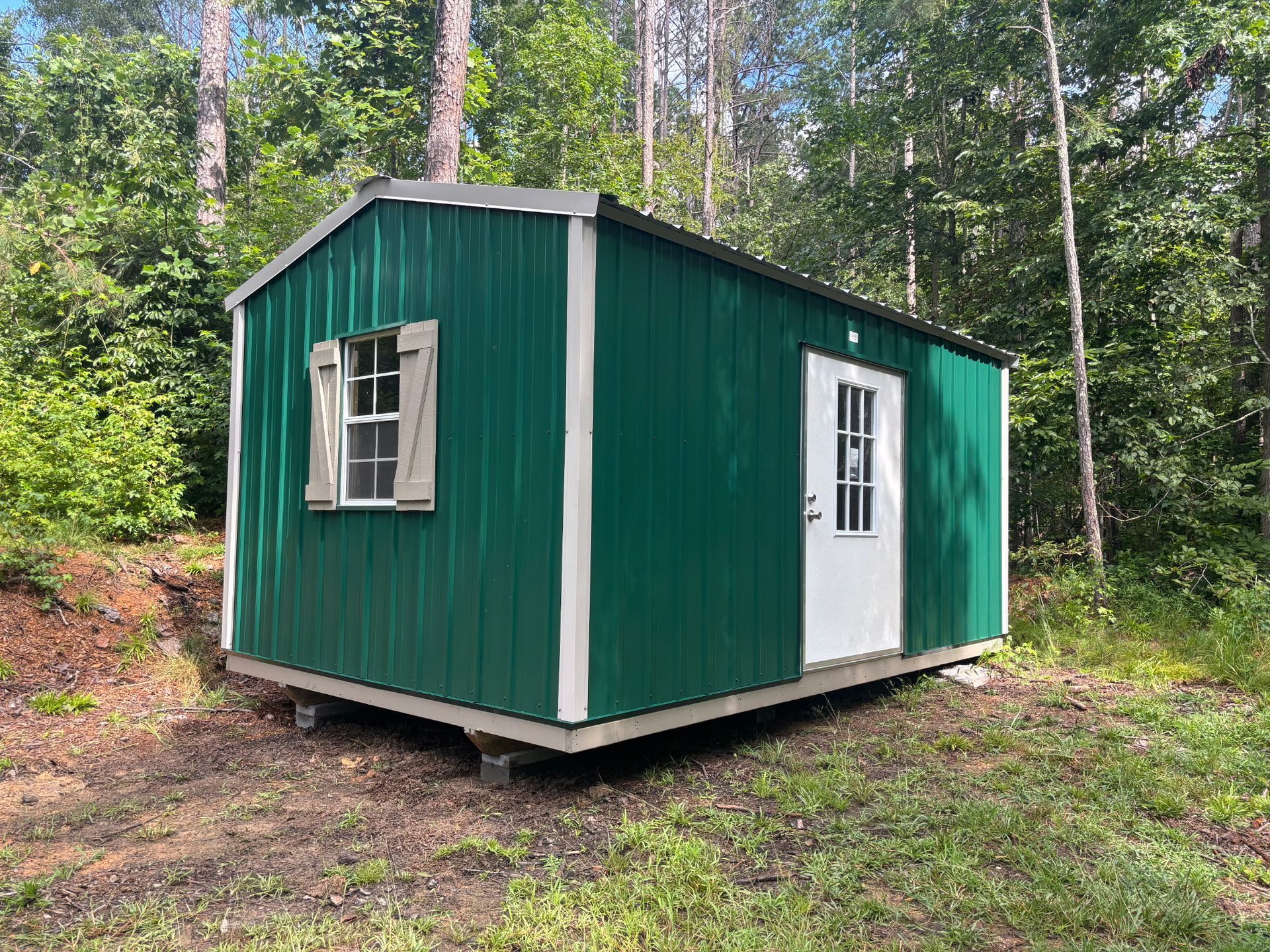 Green metal shed with white trim, window, and door in a wooded area.