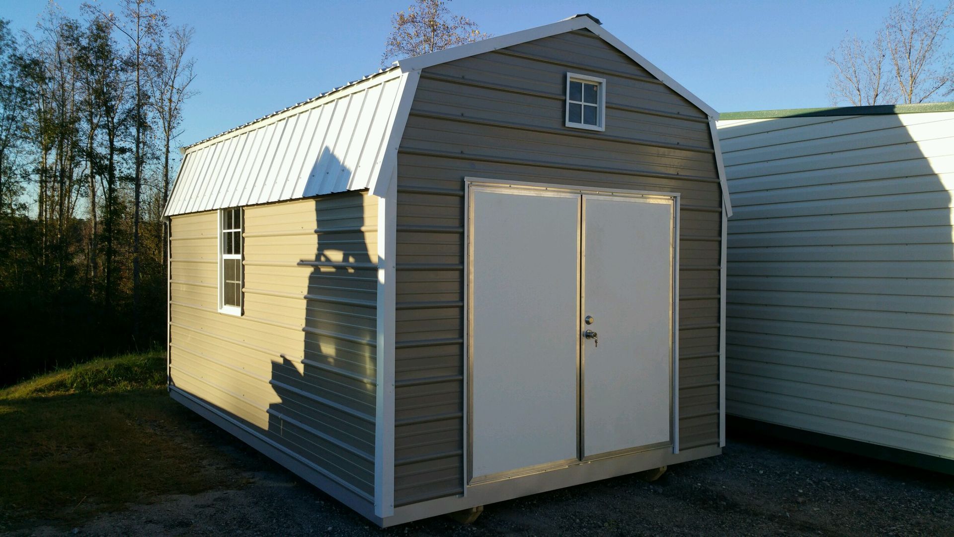 Gray shed with white doors, roof, and trim, on a grassy area, under a blue sky.