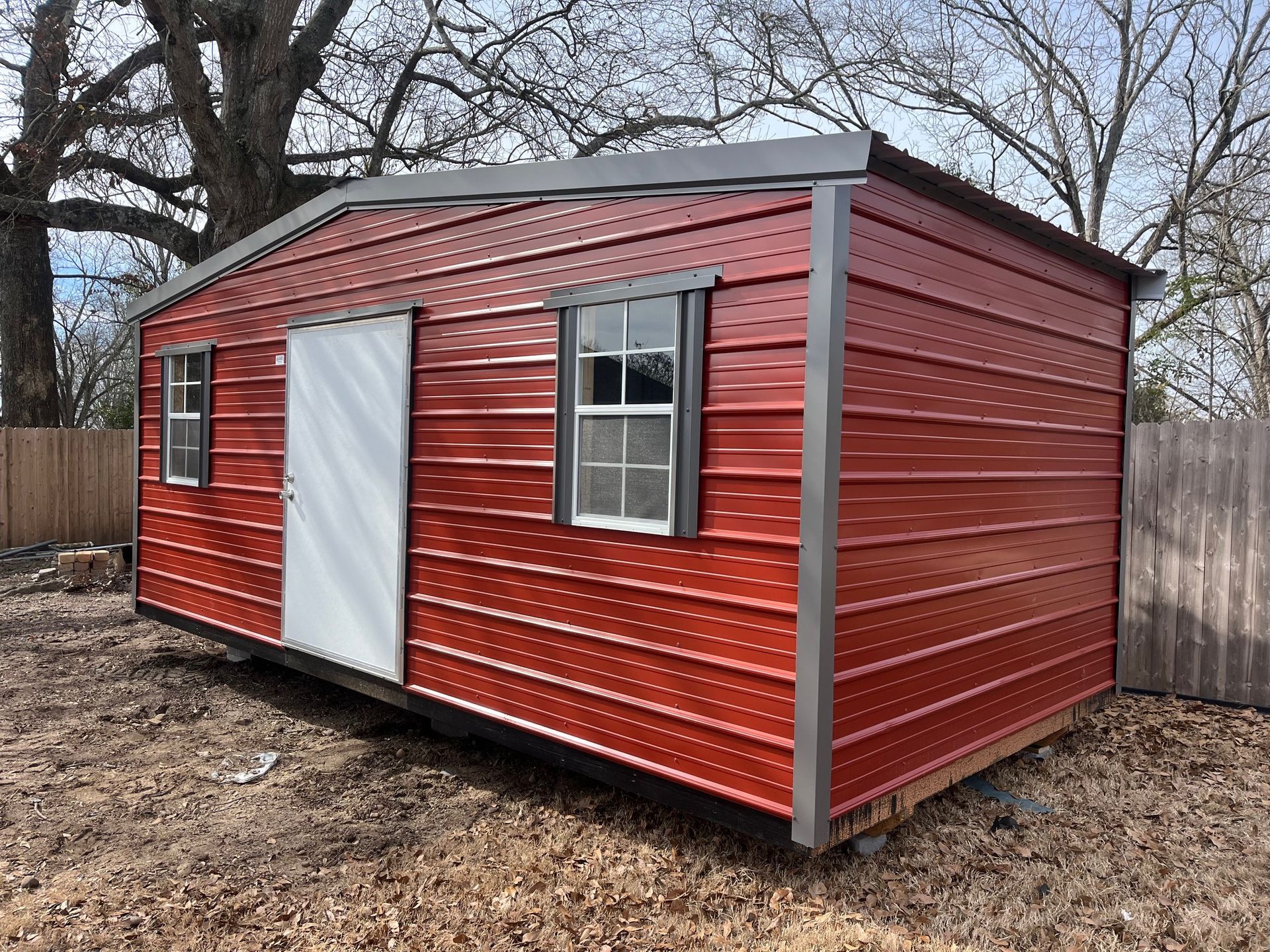 Red metal shed with white door and two windows sits on a grassy area, in front of a wooden fence.