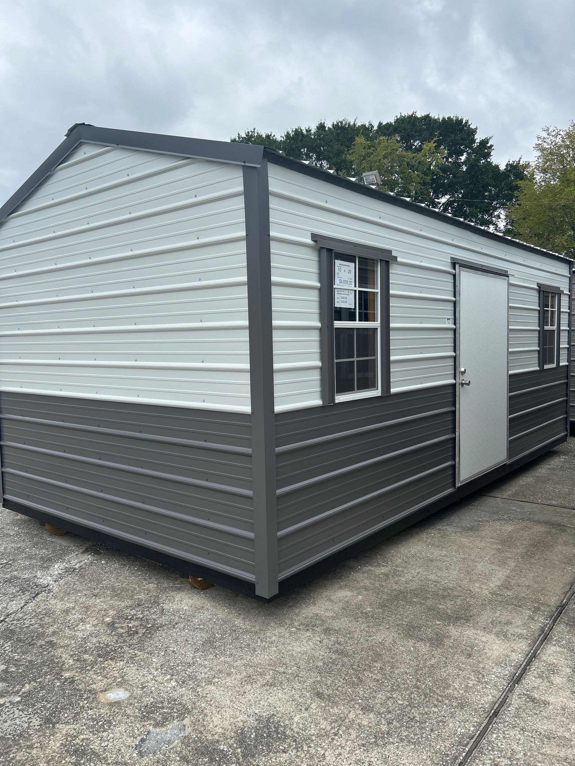 Metal storage shed, grey and white, with a door and windows, sitting on a concrete surface.