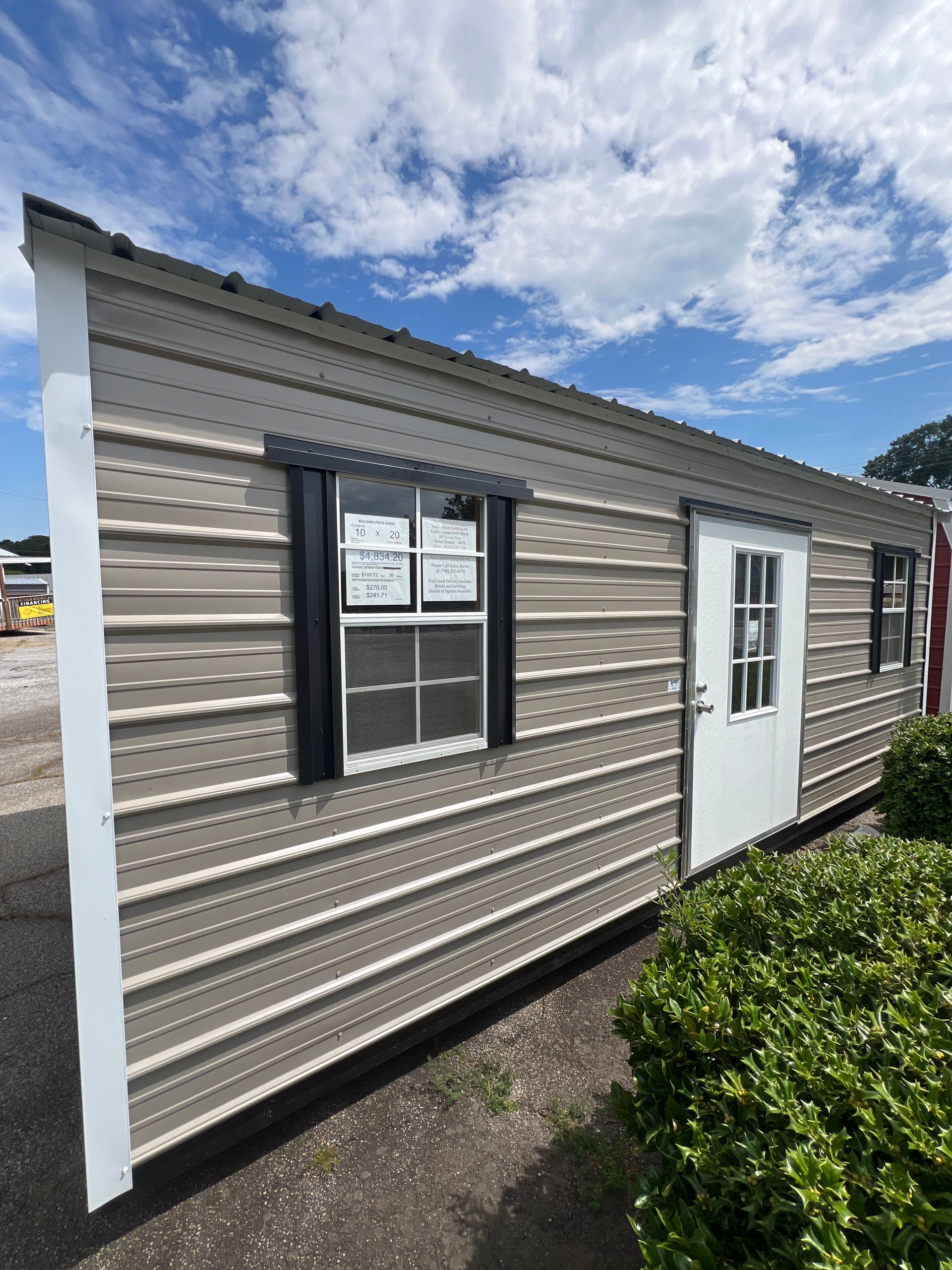 Tan-sided shed with white door and window, black window trim, in front of green bushes, under a blue sky.