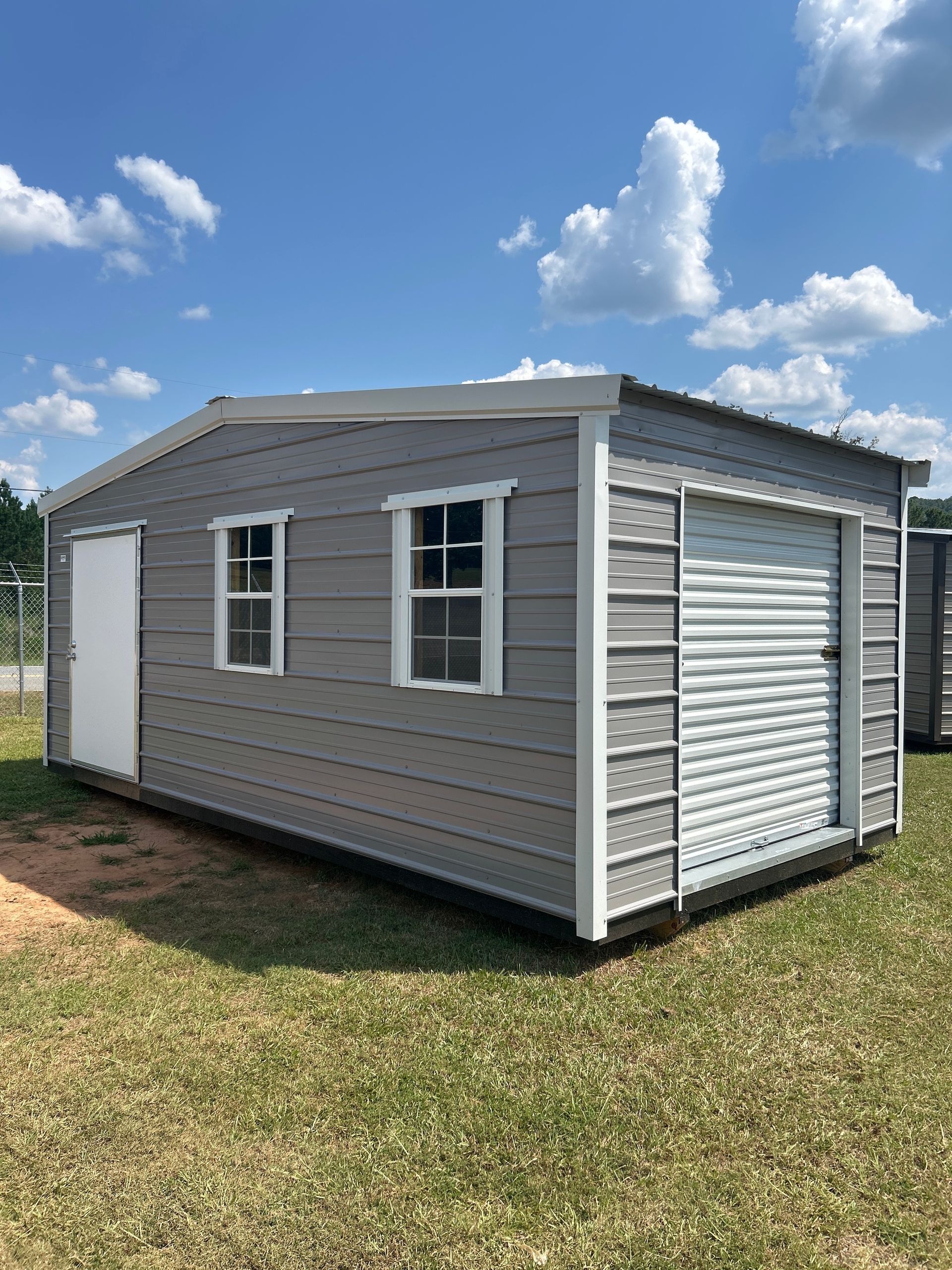 Gray shed with white trim, door, windows, and roll-up garage door on a grassy area under a blue sky.