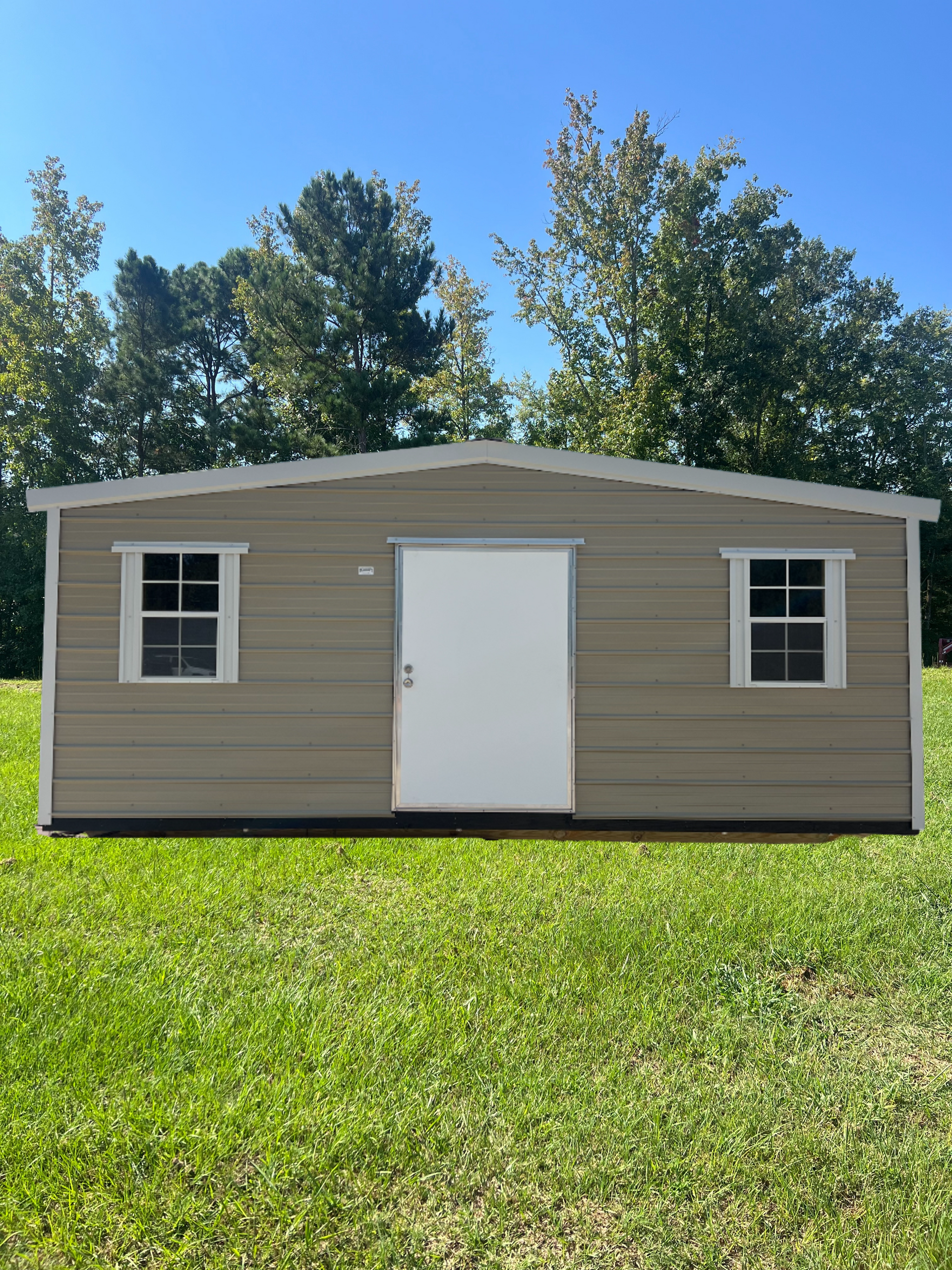Tan metal shed with white door and windows, set on green grass against a background of trees and a blue sky.