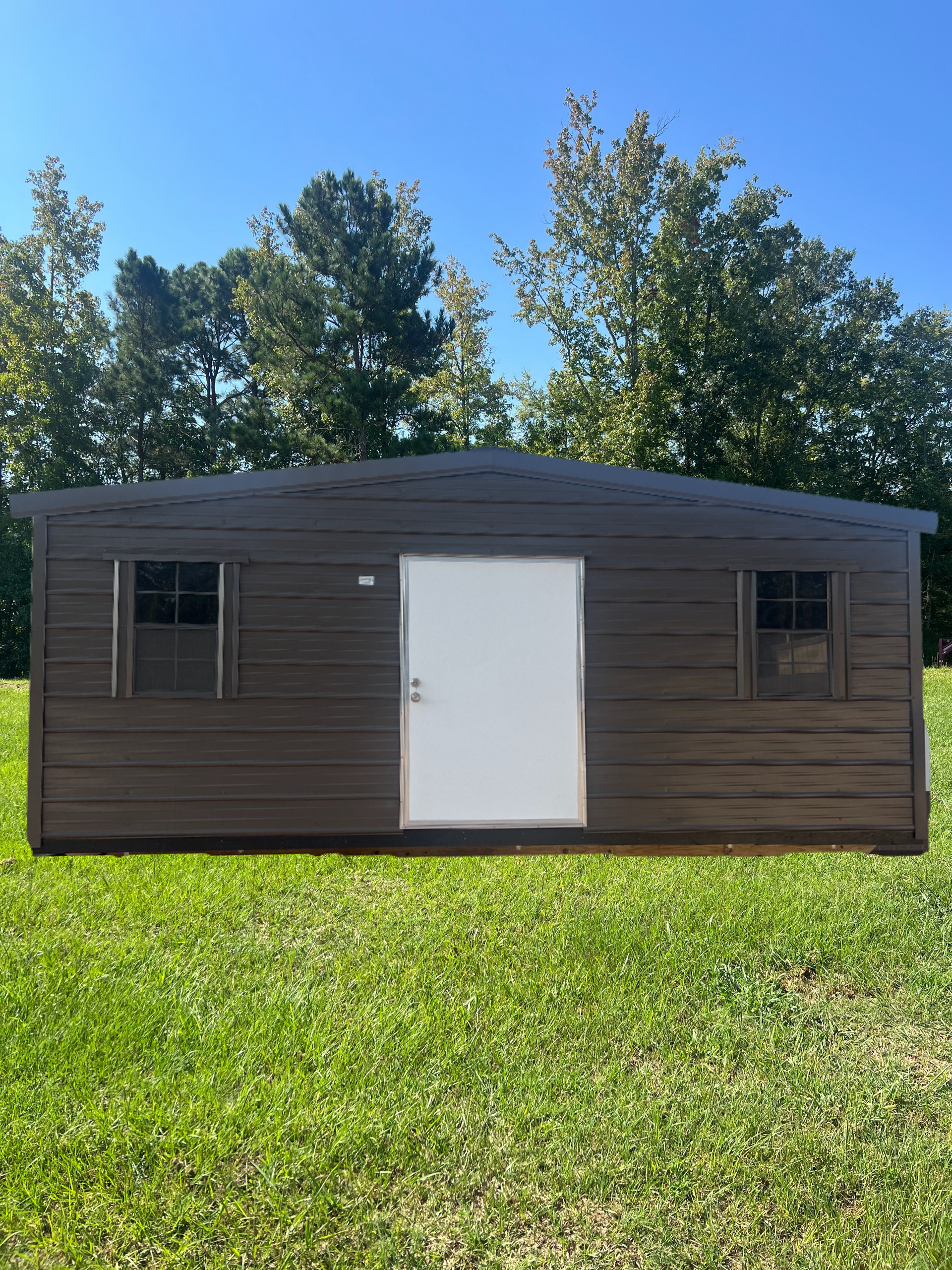 Brown shed with white door and two small windows on green grass, trees in background.