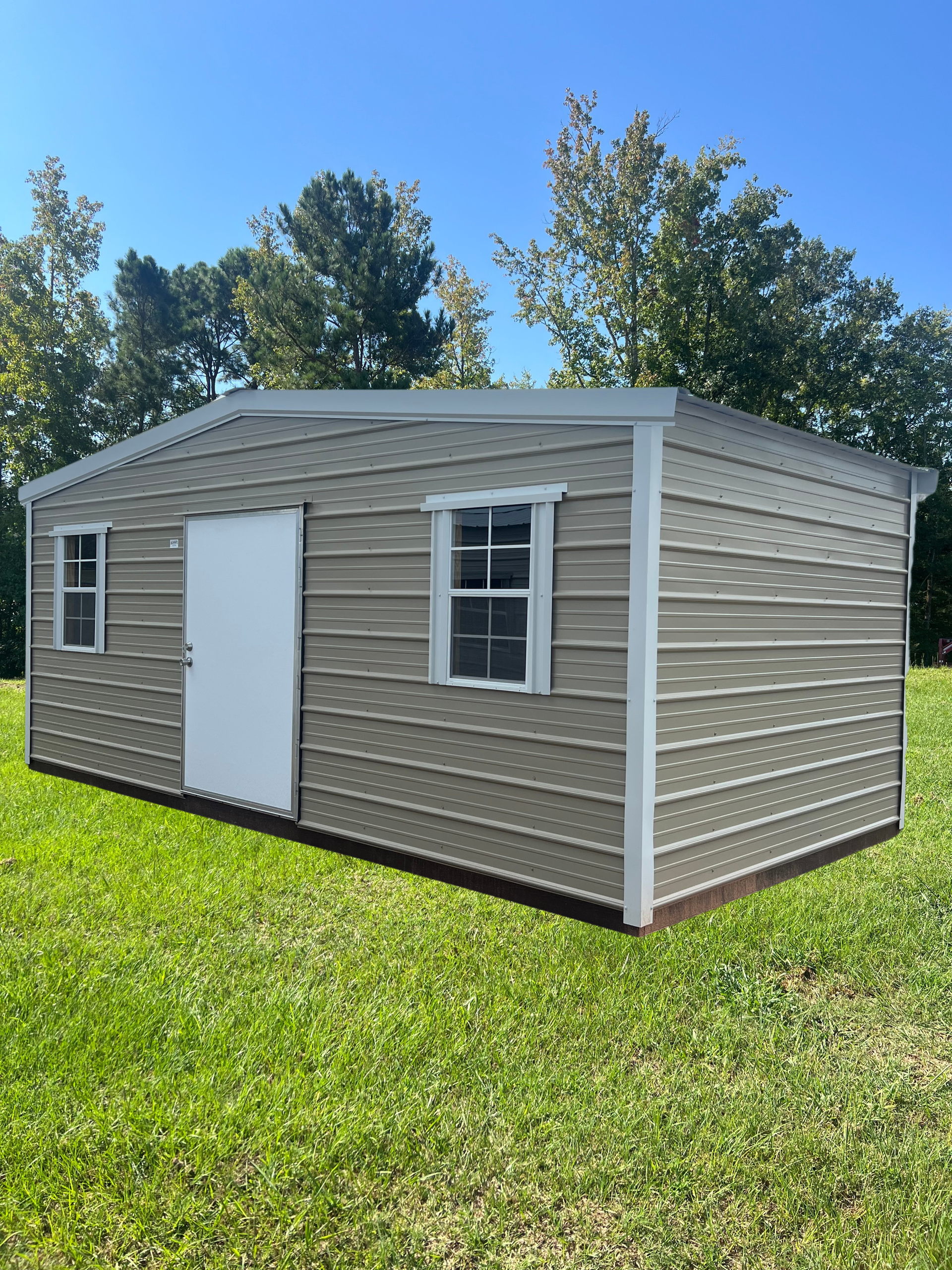 Tan metal storage shed with white door and two windows sitting on a grassy lawn under a clear blue sky.