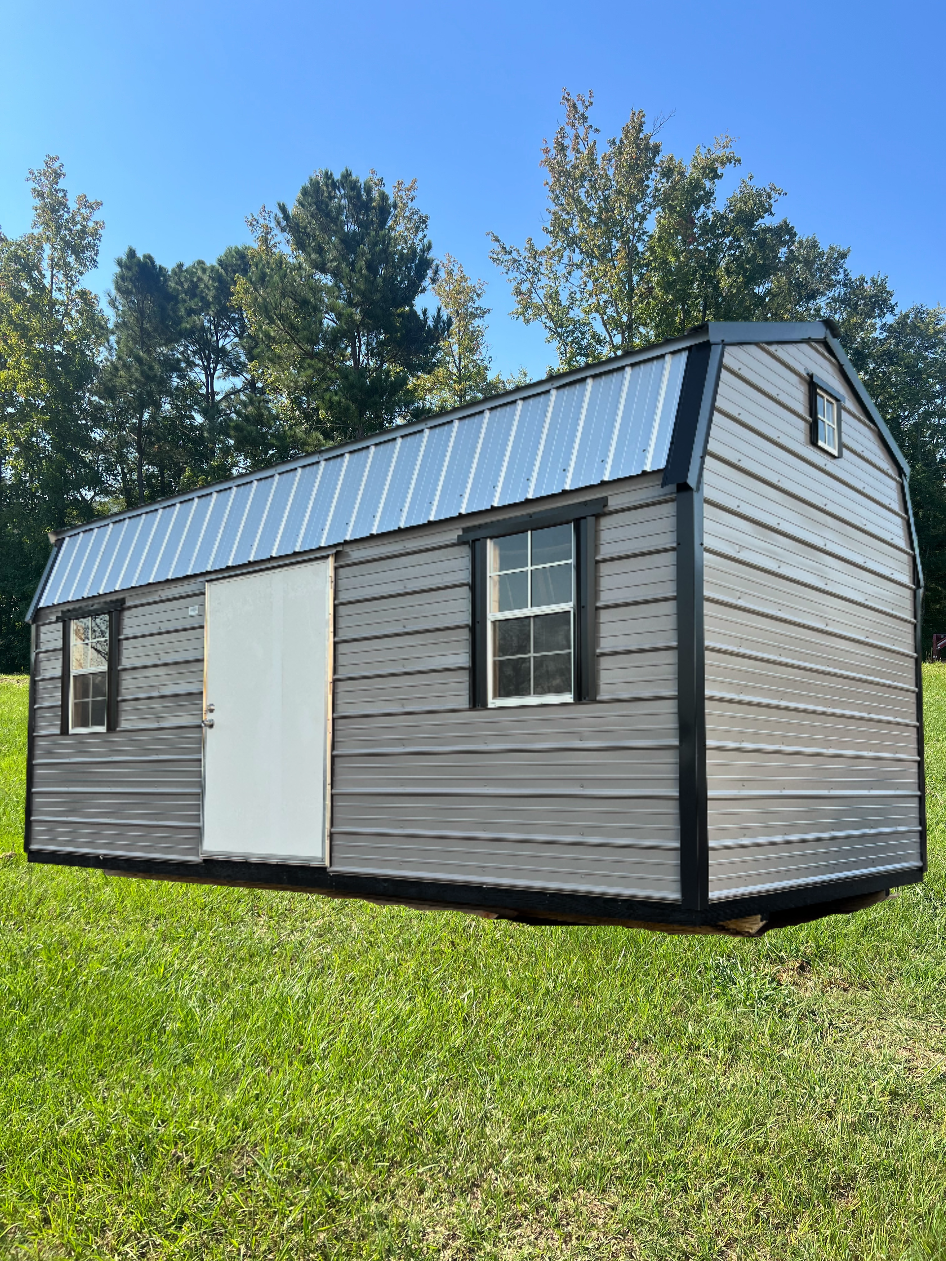 Gray and white barn-style shed with a metal roof and two windows set on a grassy lawn under a clear sky.