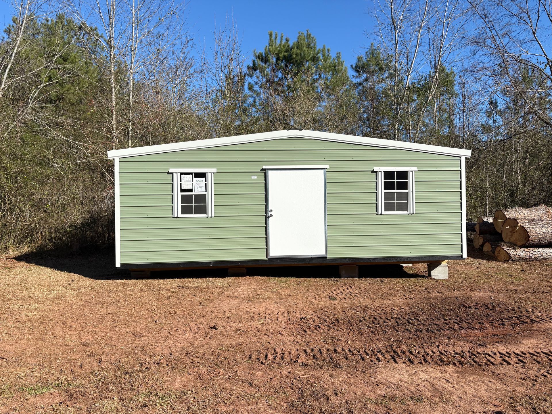 Green metal building with a white door and two windows, set in a wooded area on a sunny day.