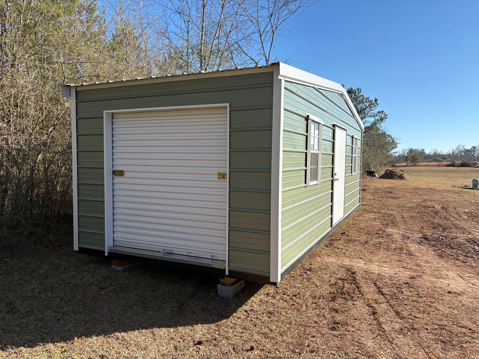 Green metal shed with white garage door, small windows, and a door on a sunny day.