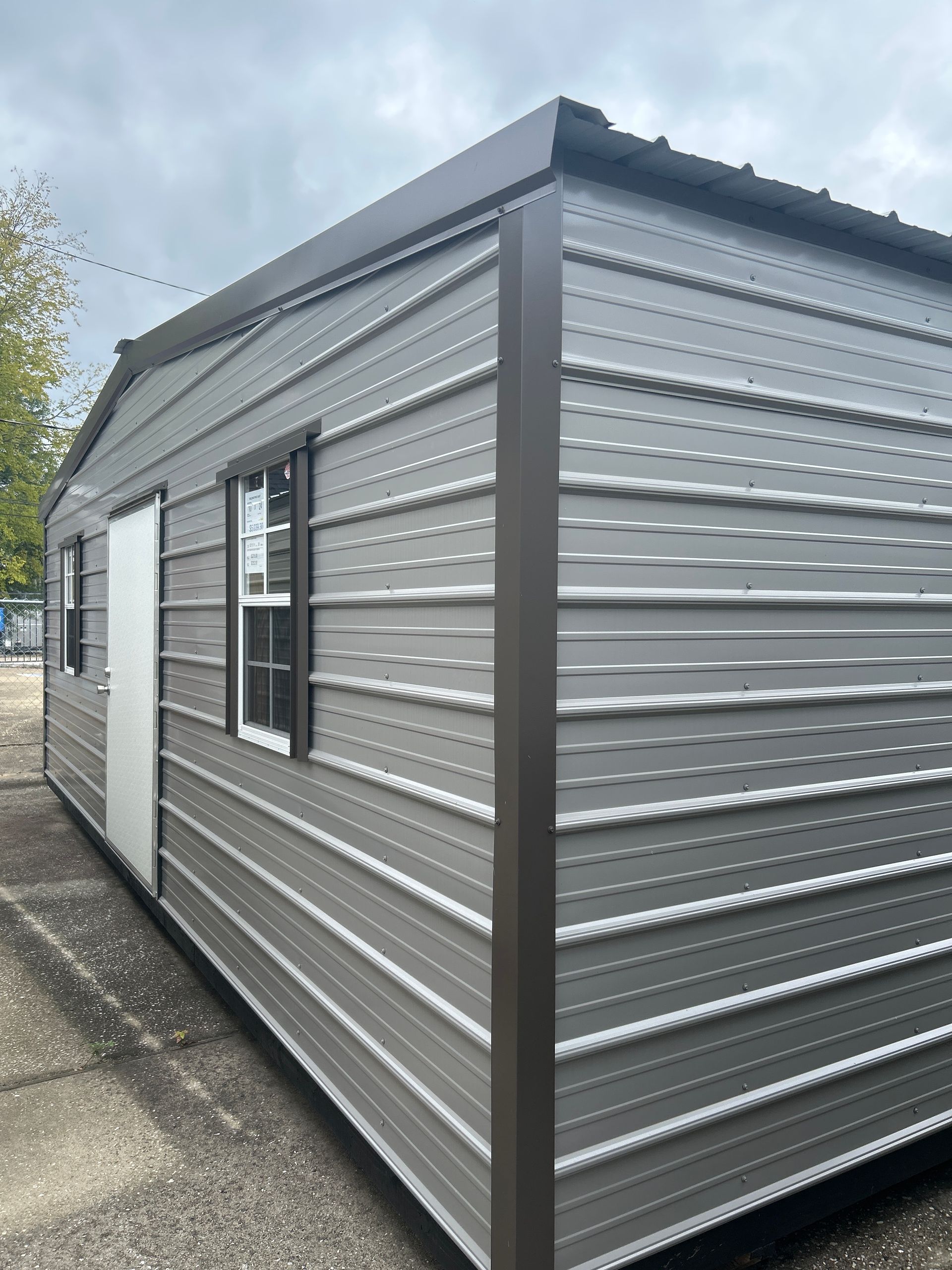 Gray metal shed with door and windows, on a paved surface, overcast sky.
