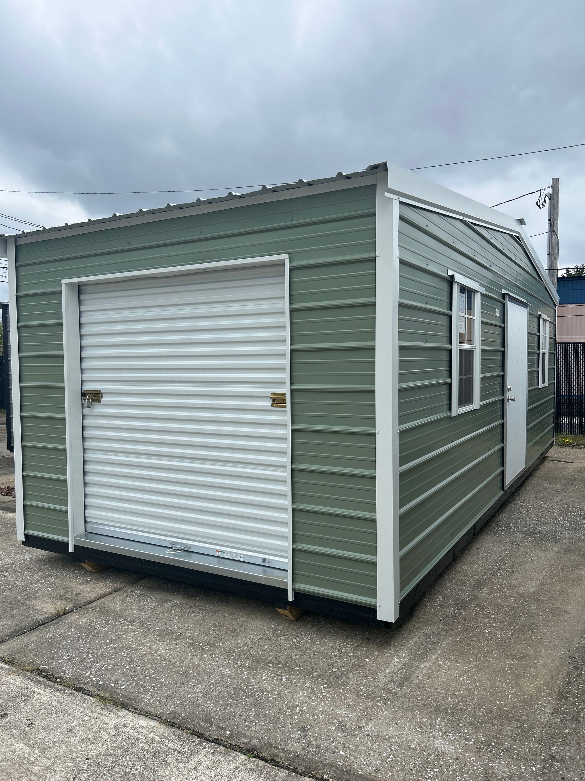 Green metal storage shed with a roll-up door and small windows, on a concrete surface.