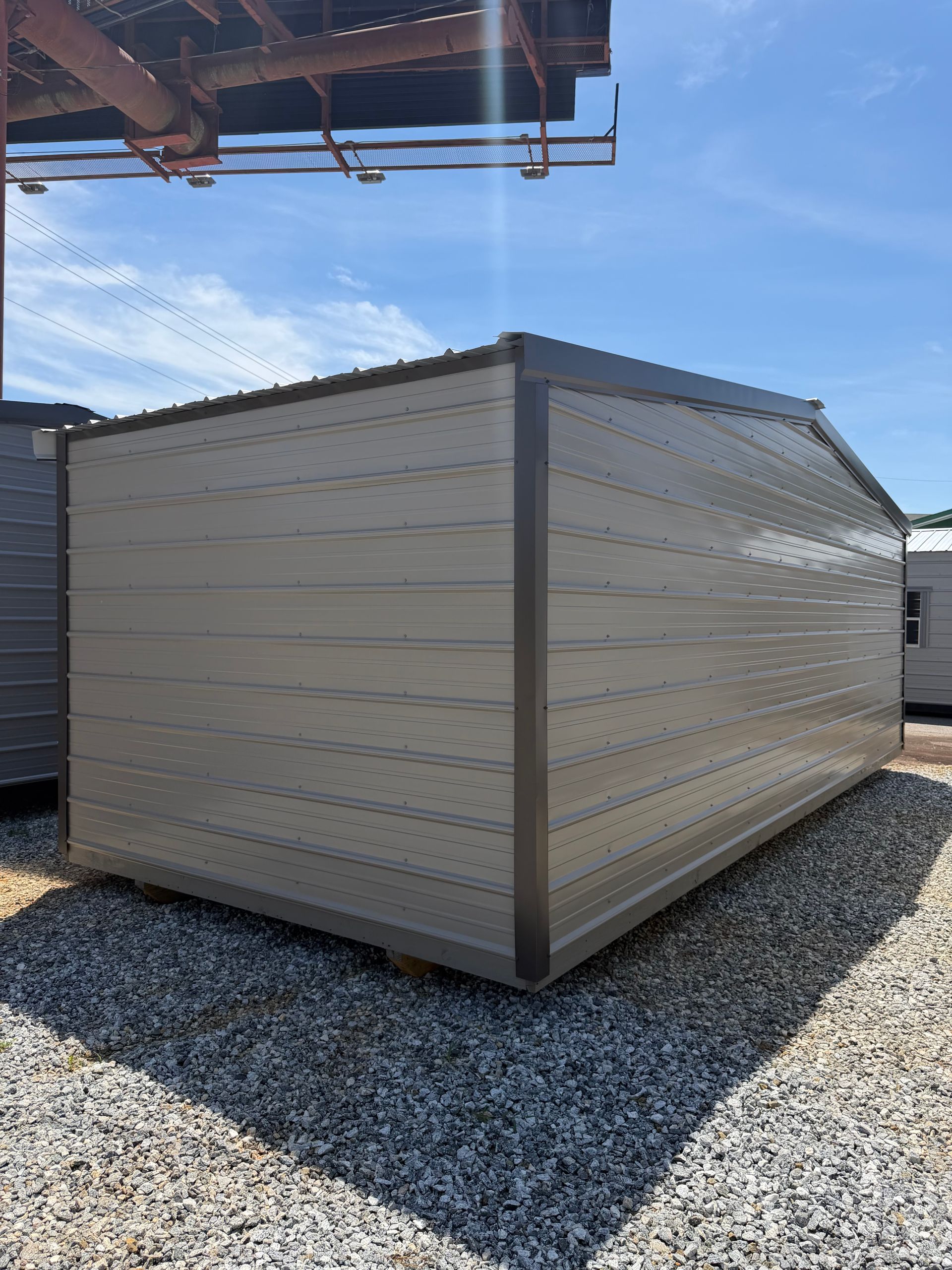 Beige metal storage shed on gravel beside a building under a sunny blue sky