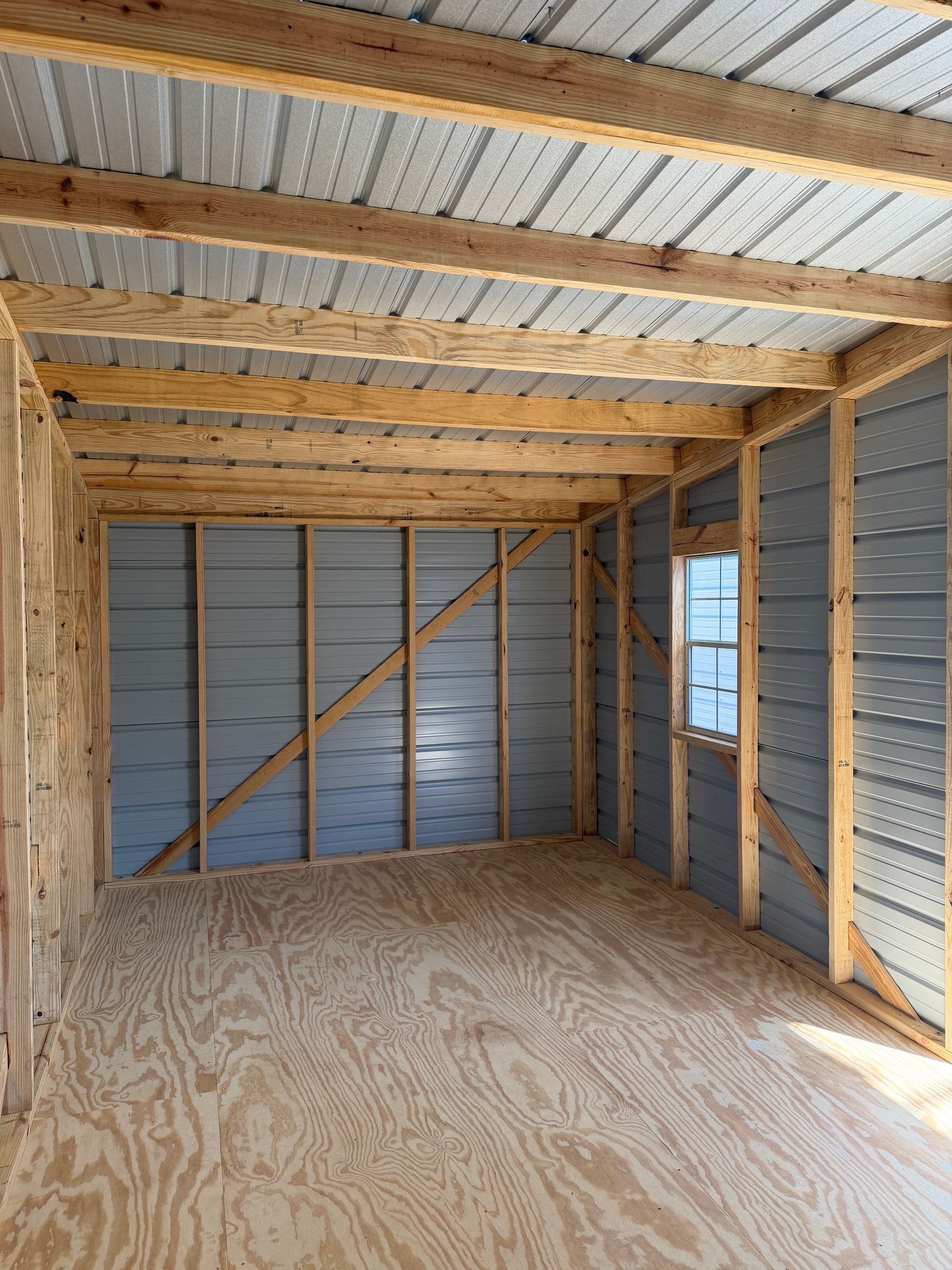 Empty unfinished shed interior with plywood floor, exposed wood framing, and corrugated metal walls and ceiling.