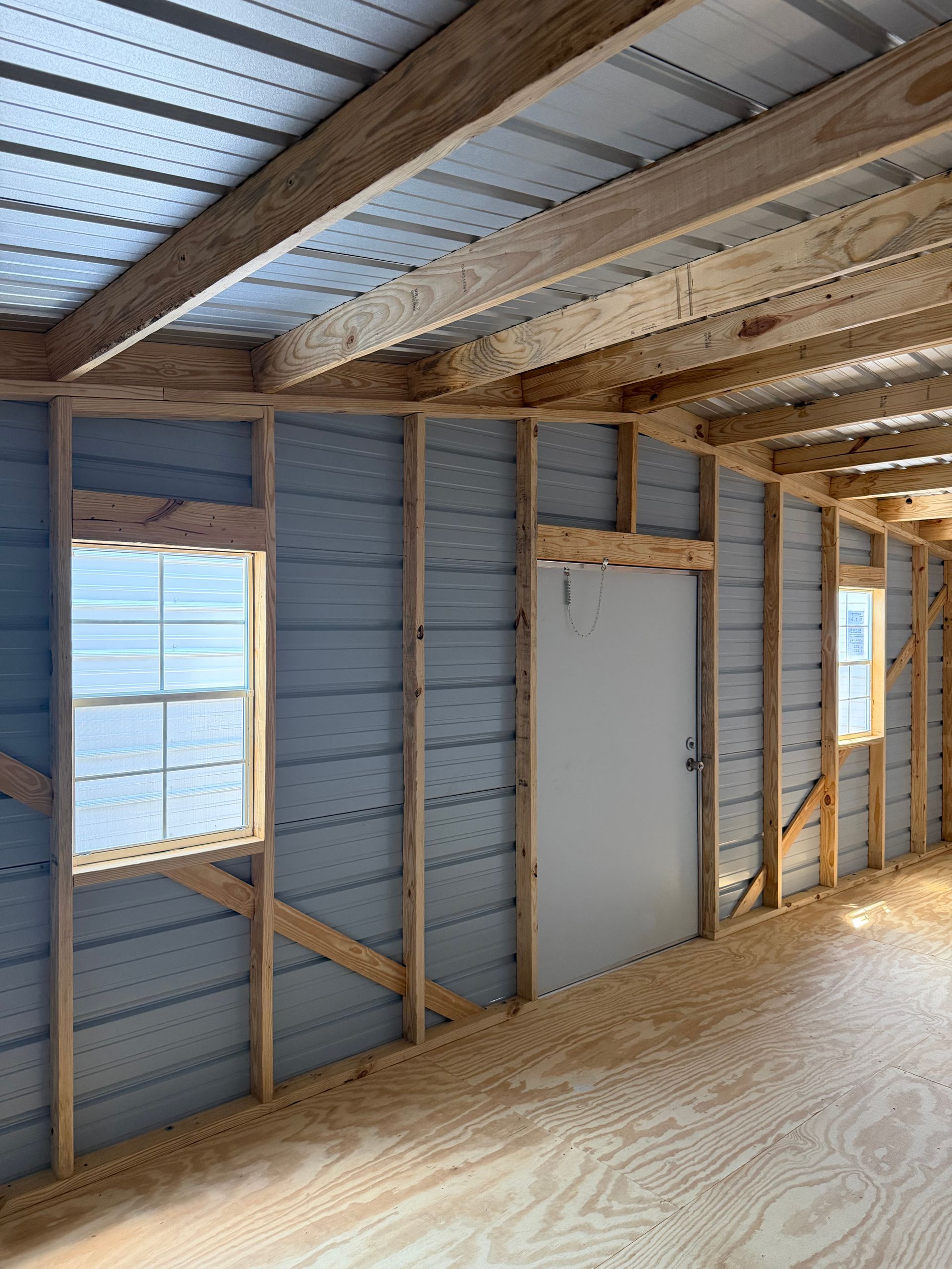Unfinished interior with exposed wood framing, corrugated metal ceiling, and two windows beside a white door.