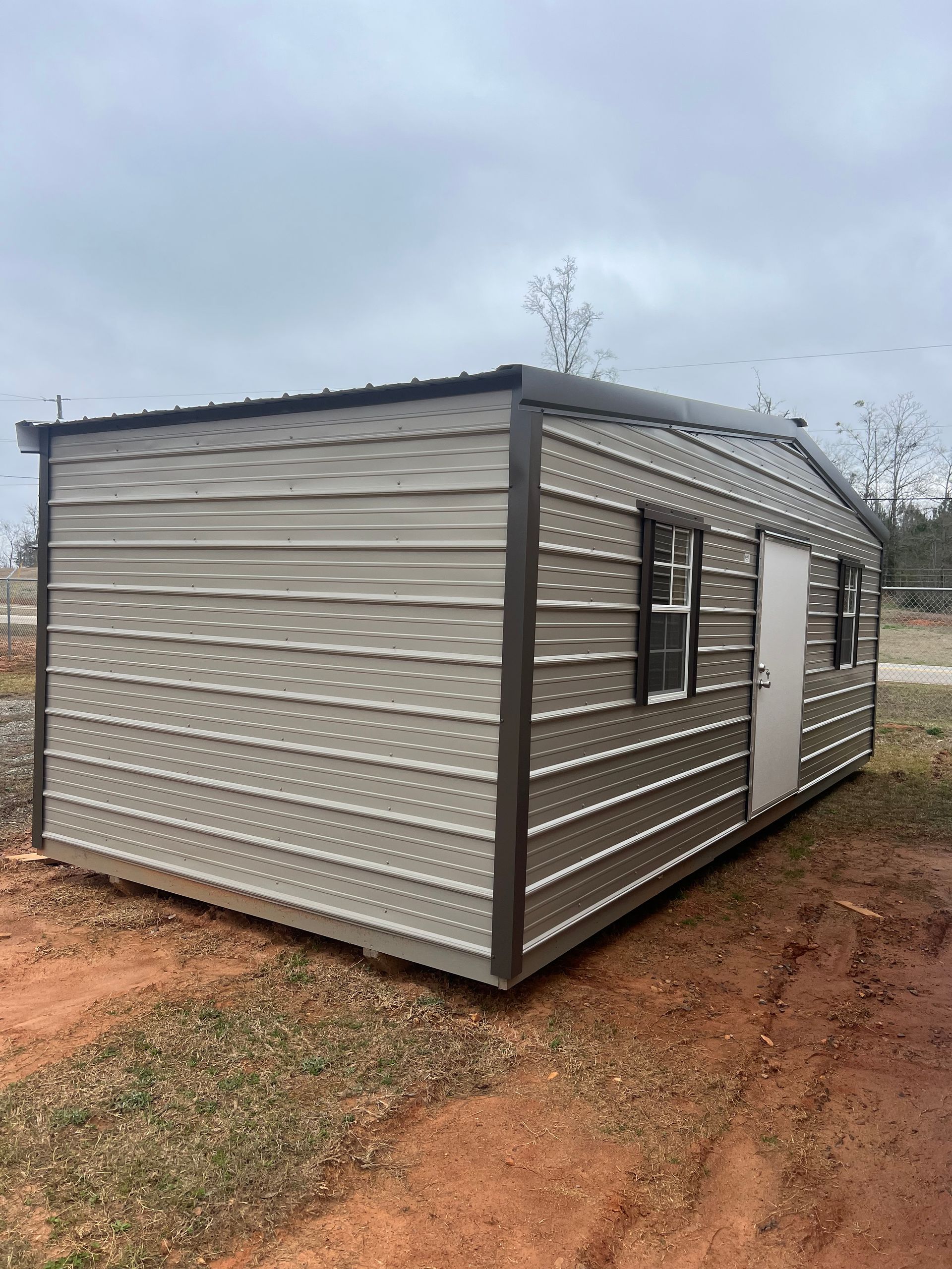 Tan metal shed with dark trim, windows, and door, set on a grassy area.
