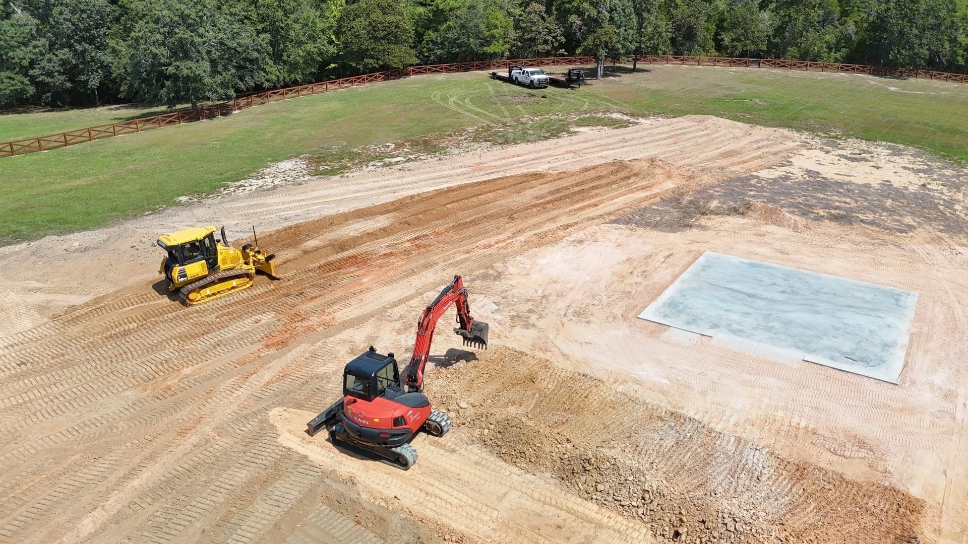 Construction site with bulldozer and excavator leveling soil near a concrete pad.