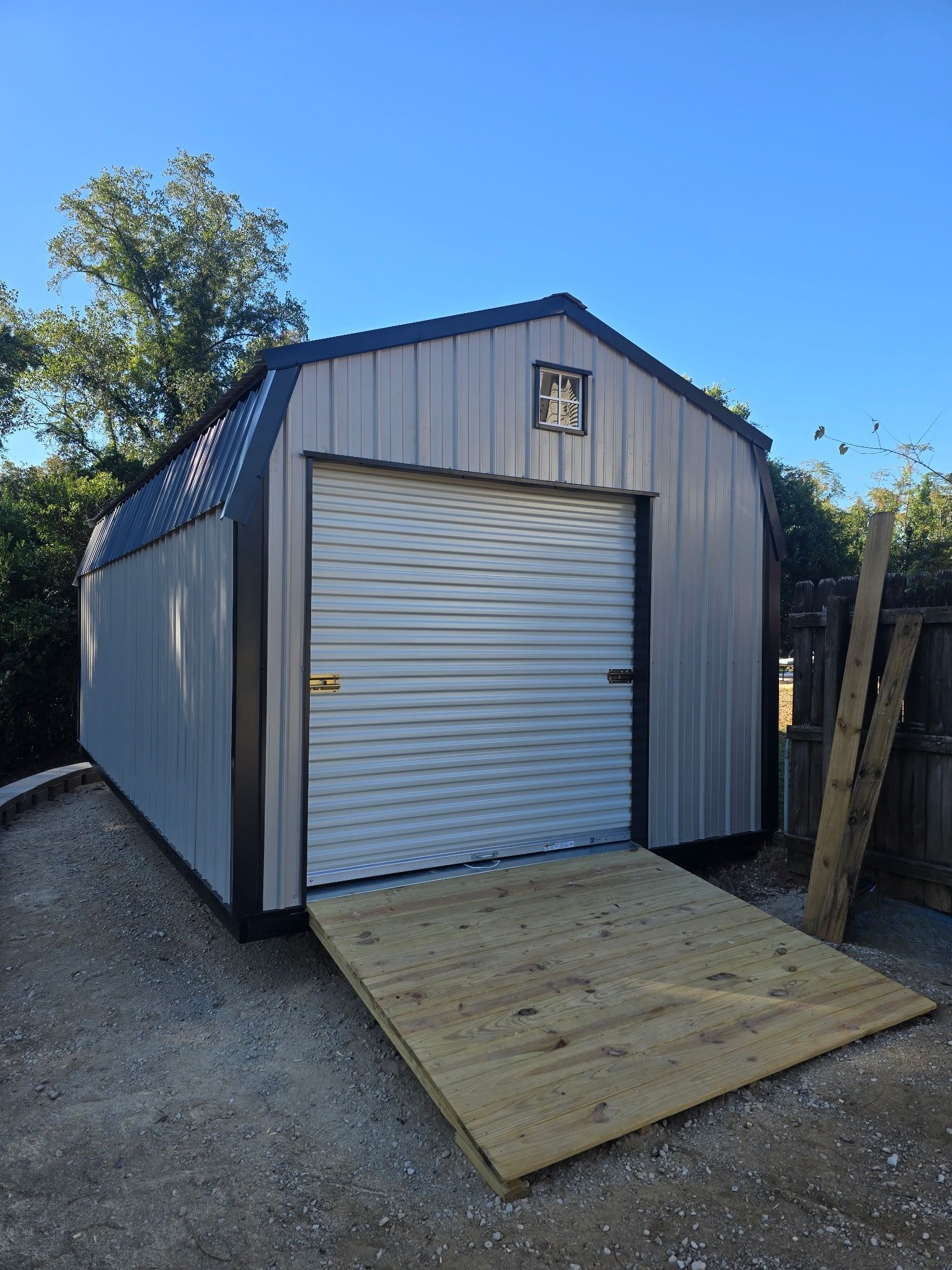Gray metal shed with a roll-up door, wooden ramp, and a blue sky background.