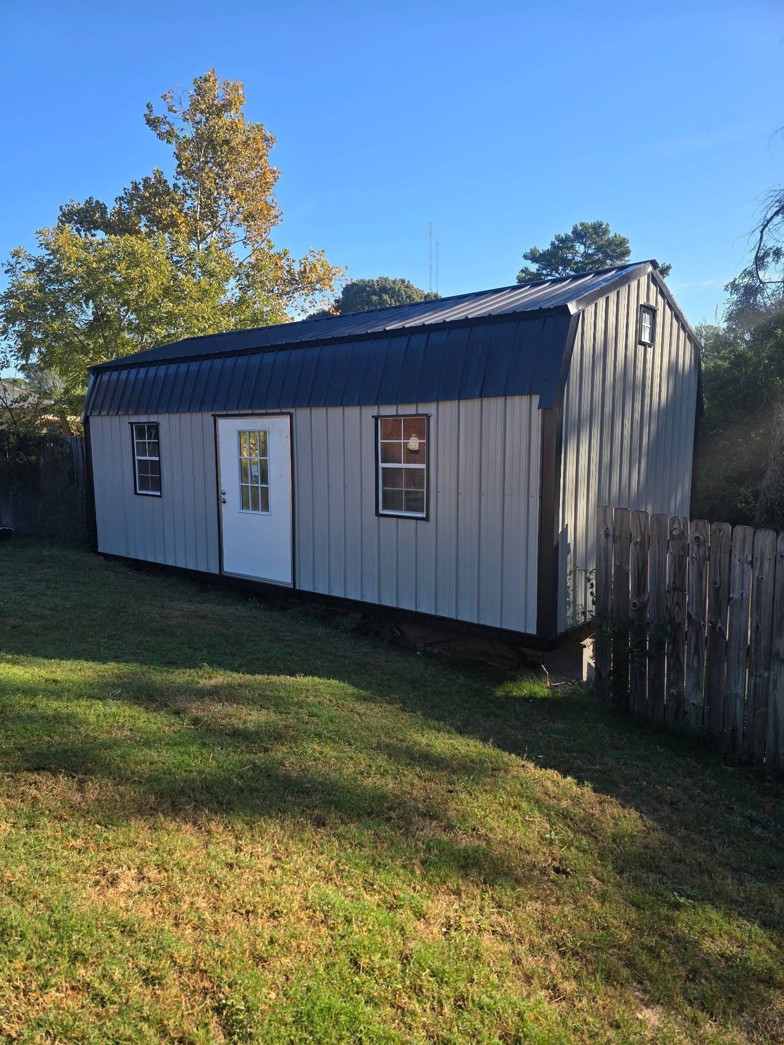 Gray shed with black roof, white door, and two windows sits on a grassy hill.