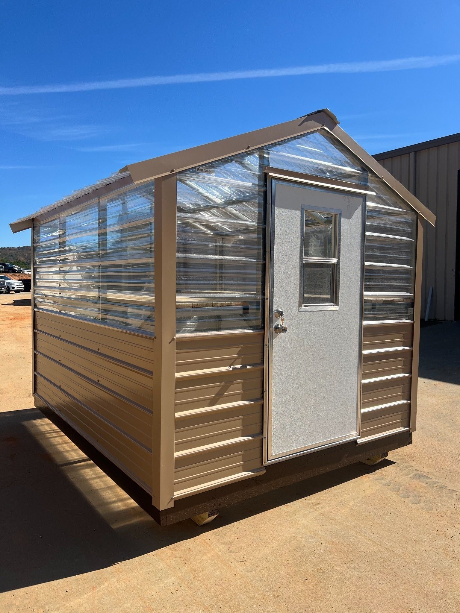 Tan and clear panel greenhouse with a white door and metal roof.
