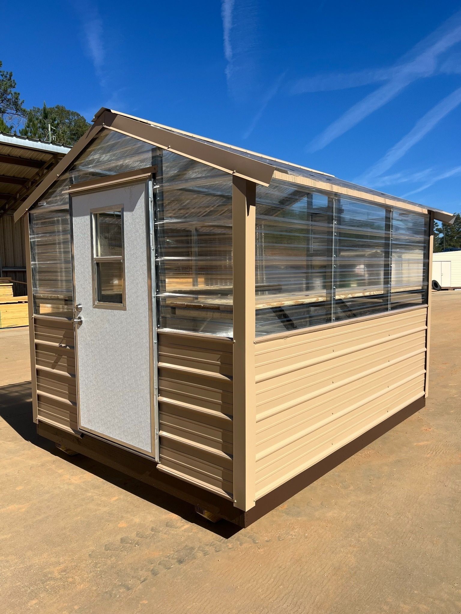 Tan and clear greenhouse with a door, brown trim, and a blue sky in the background.