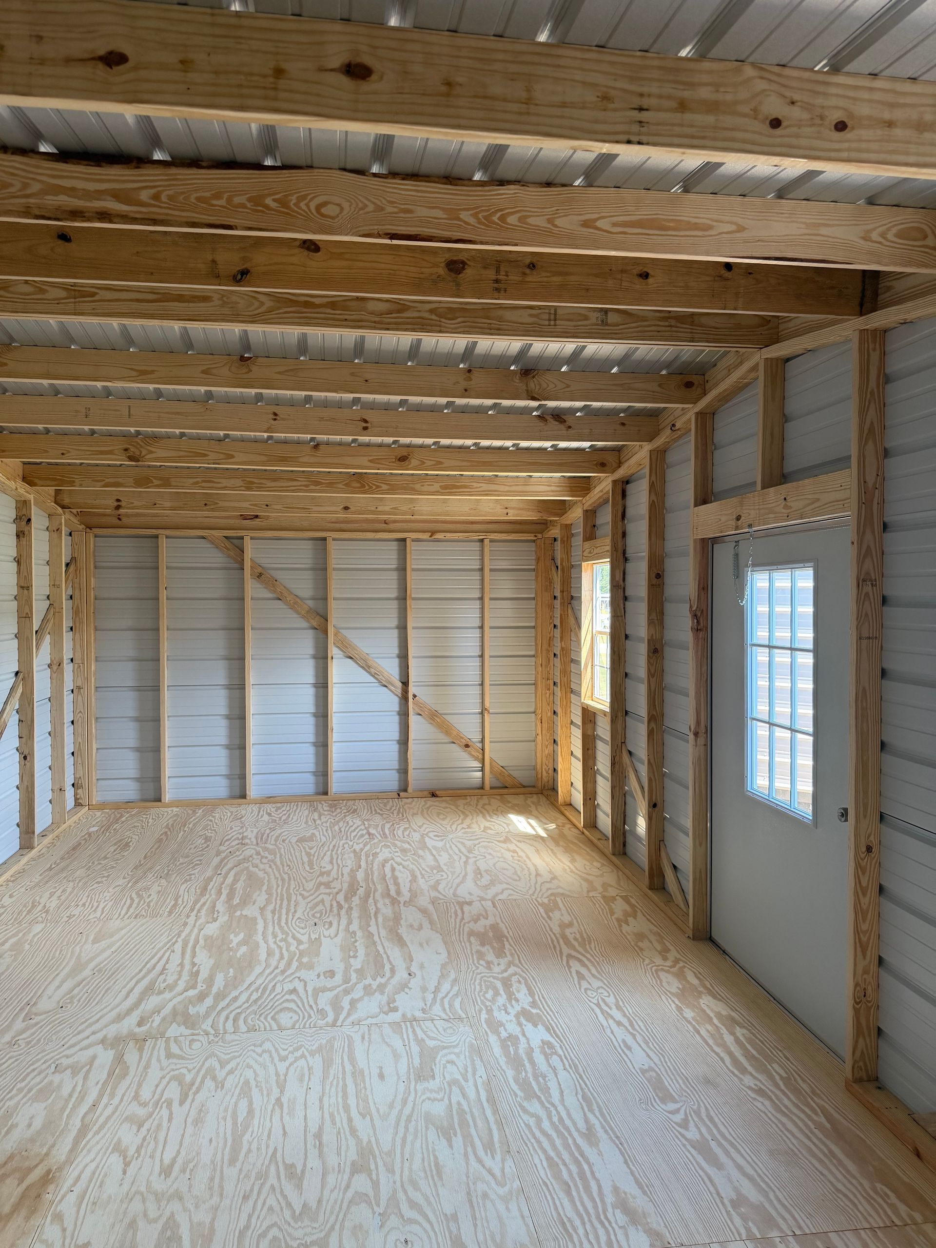 Unfinished wooden interior of a small building with exposed beams, plywood floor, and a window on the right.
