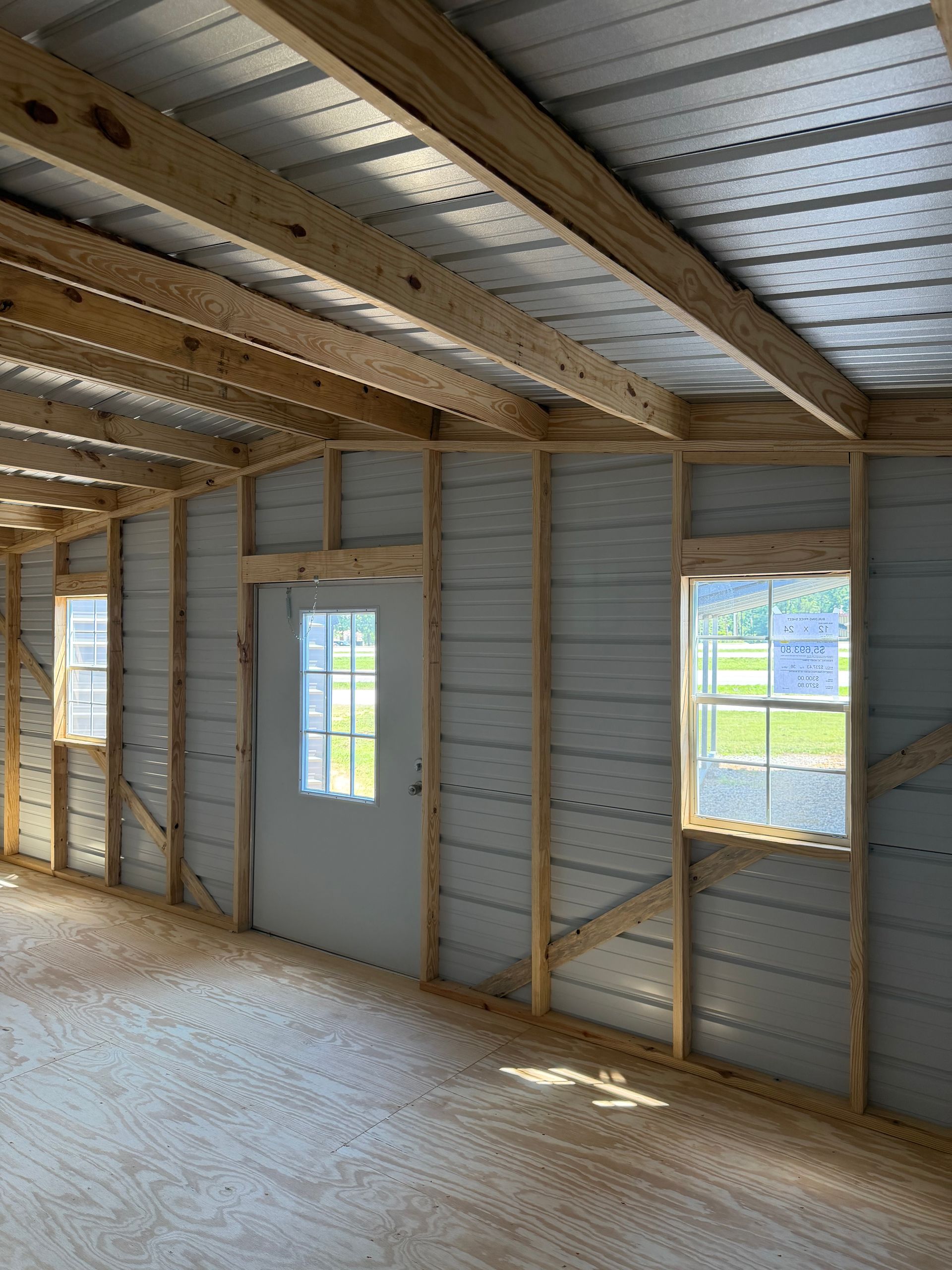 Interior of unfinished building with exposed wood framing, metal roof, and two windows along a plywood wall.