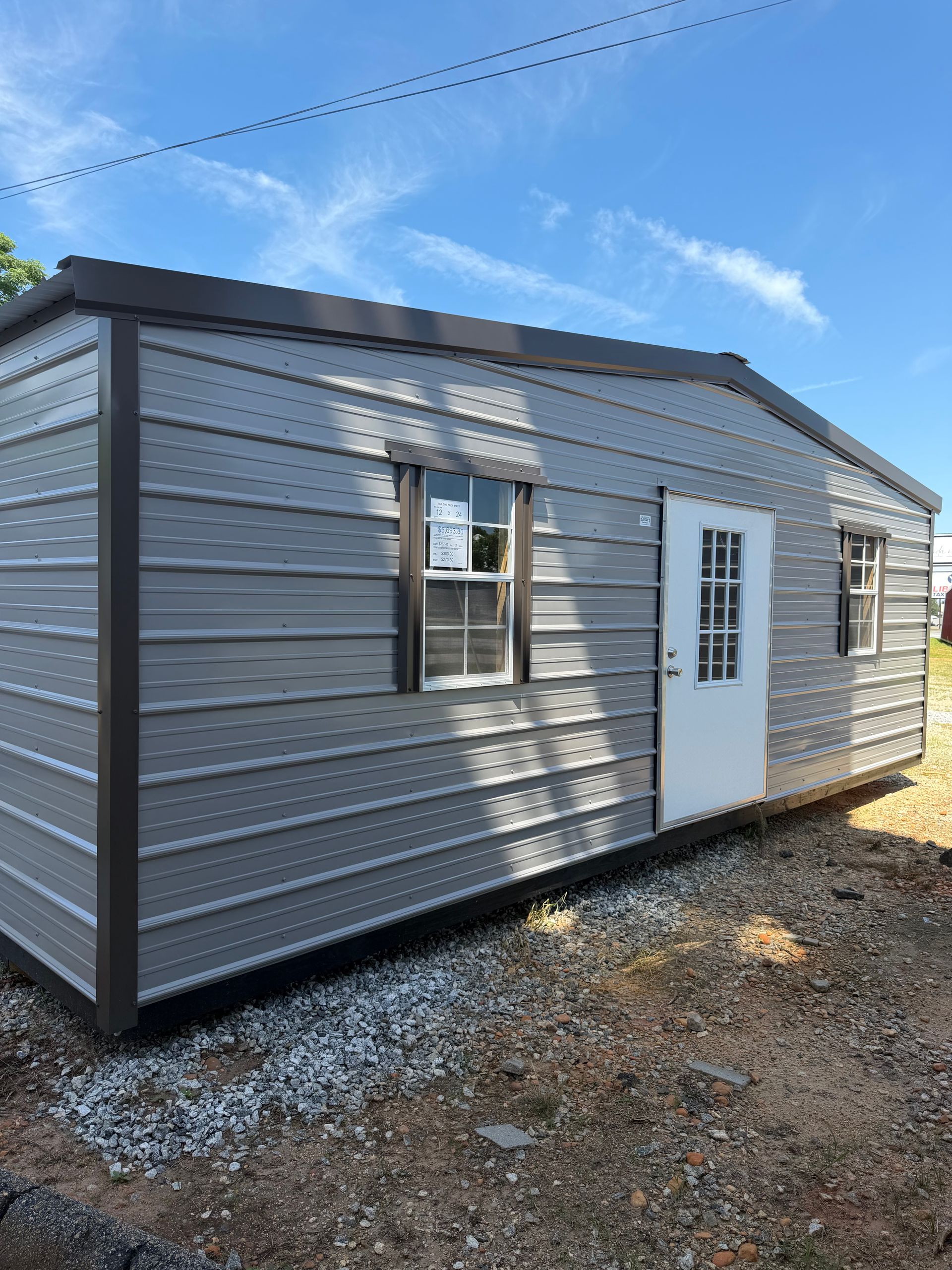 Small gray portable cabin with white door and window on a gravel lot under a blue sky