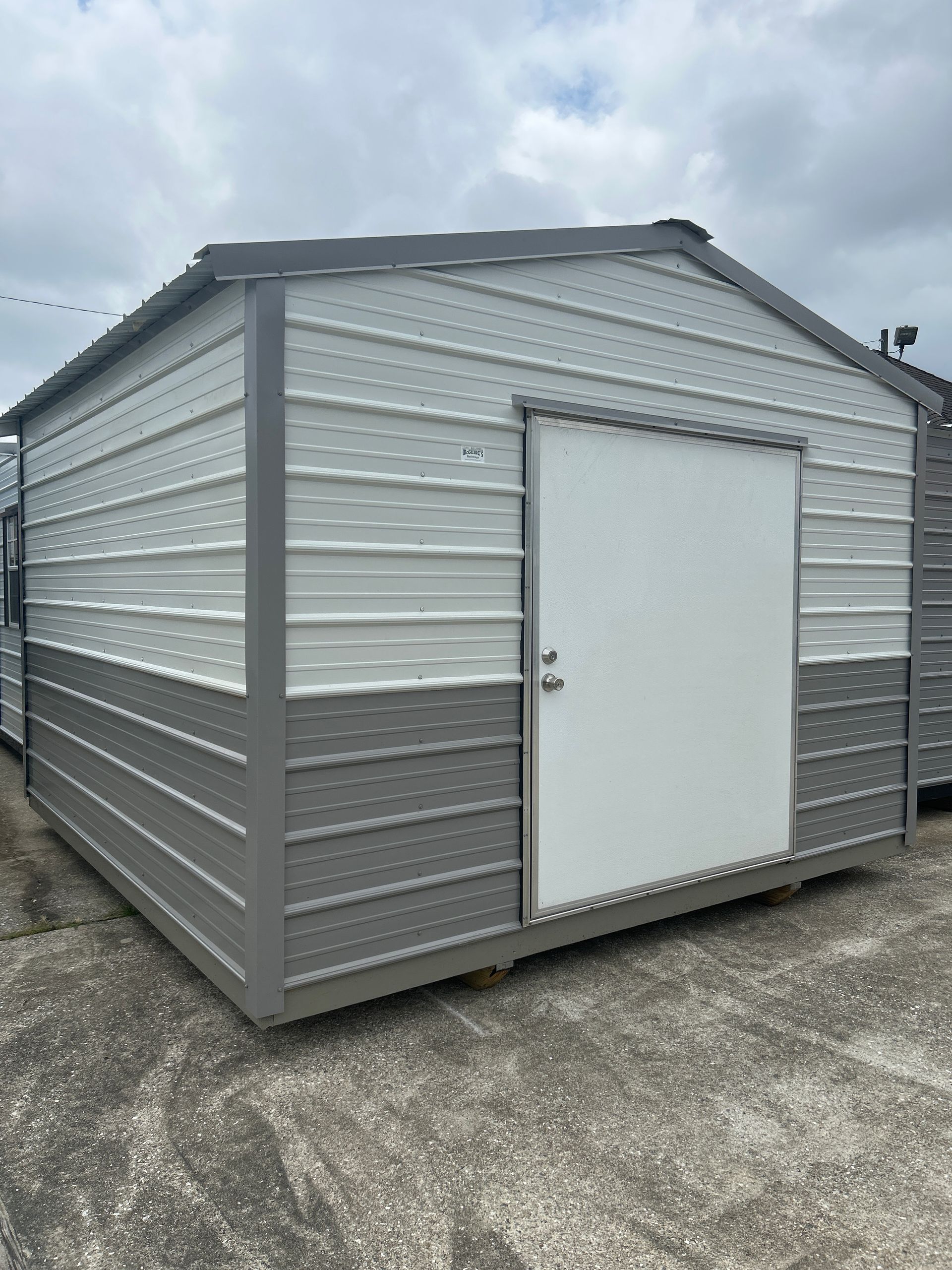 Gray and white metal storage shed with a white door, on a concrete surface, under a cloudy sky.