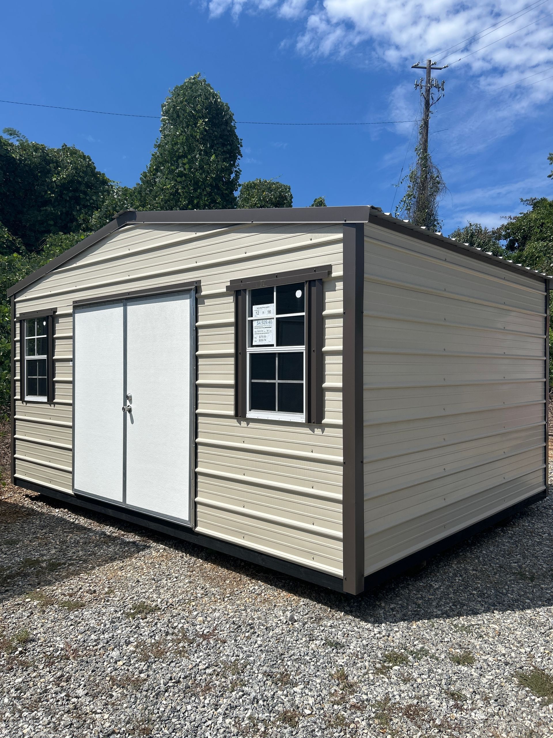 Tan metal storage shed with brown trim, double doors, two windows, gravel ground, and trees.