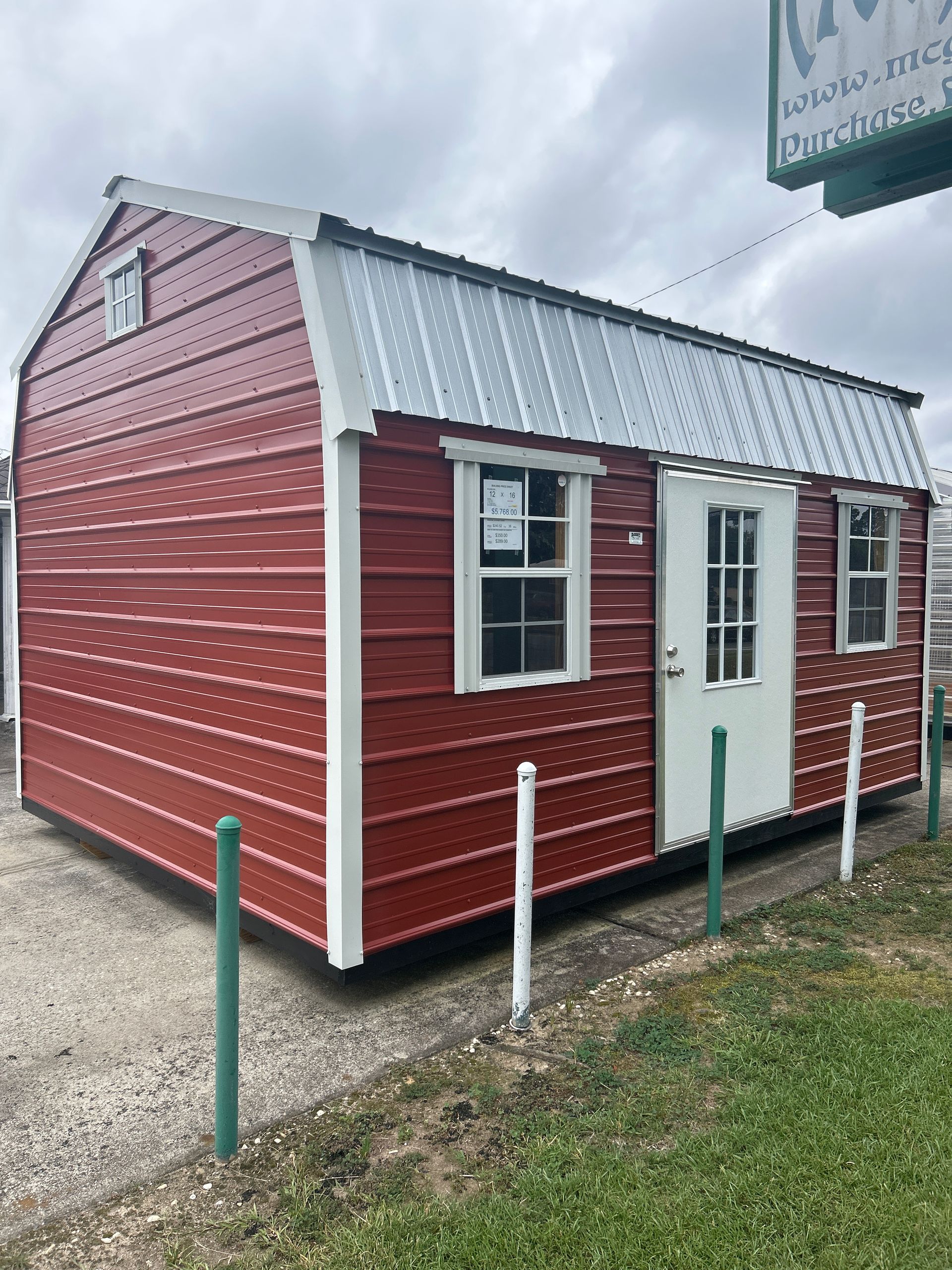 Red barn-style shed with white trim and door, metal roof, standing on a gravel lot.