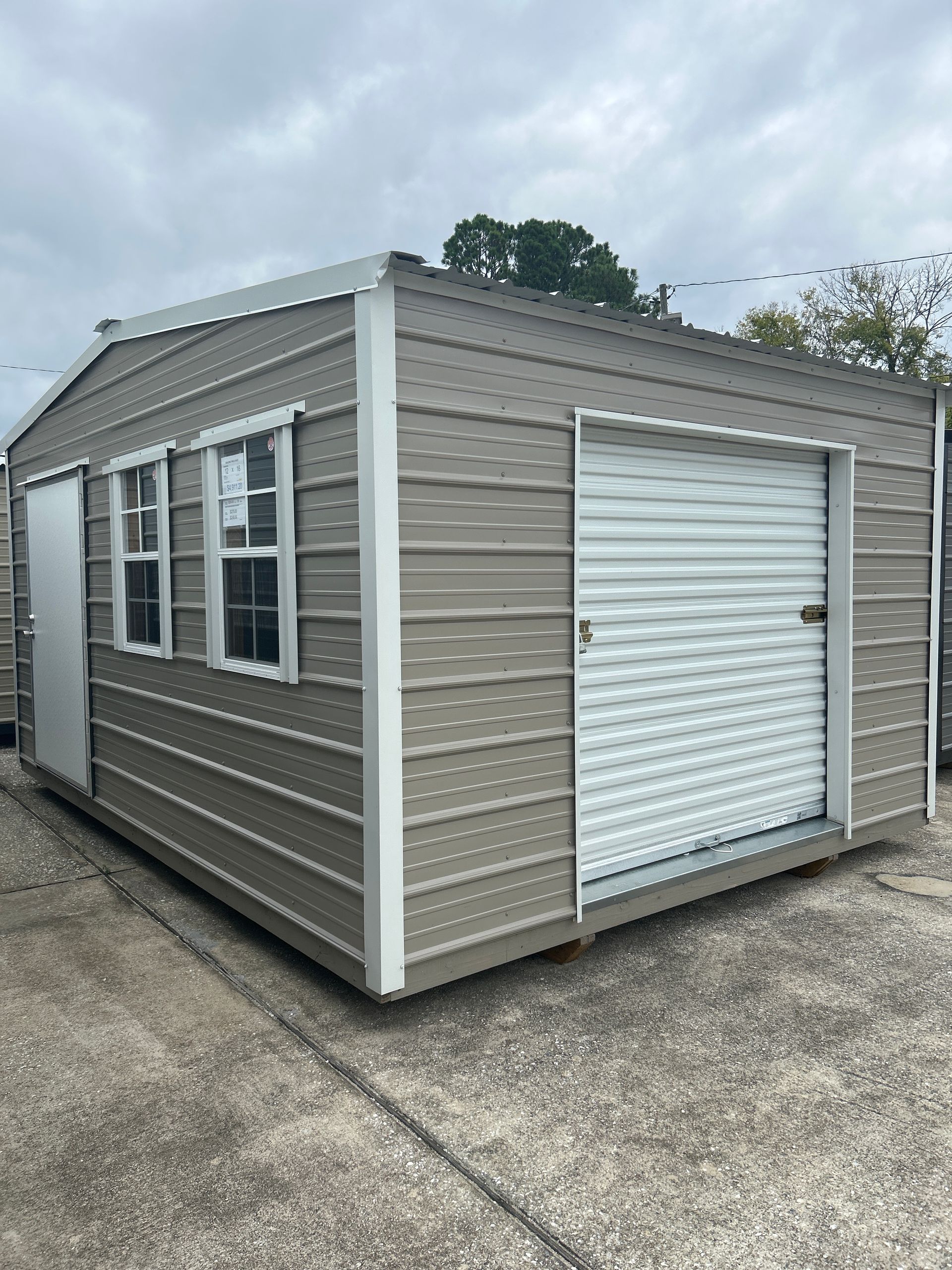 Tan metal shed with white trim, windows, door, and roll-up garage door on a concrete surface.