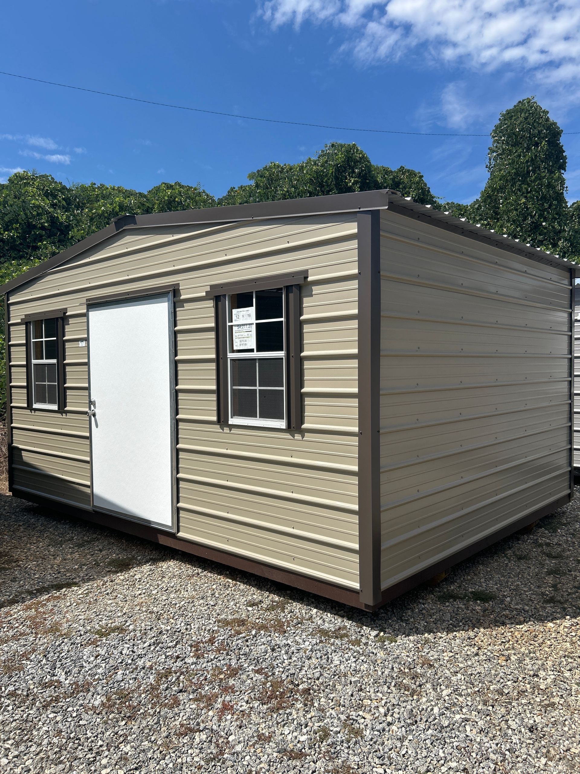 Tan metal shed with brown trim, white door, and two windows, set on gravel.