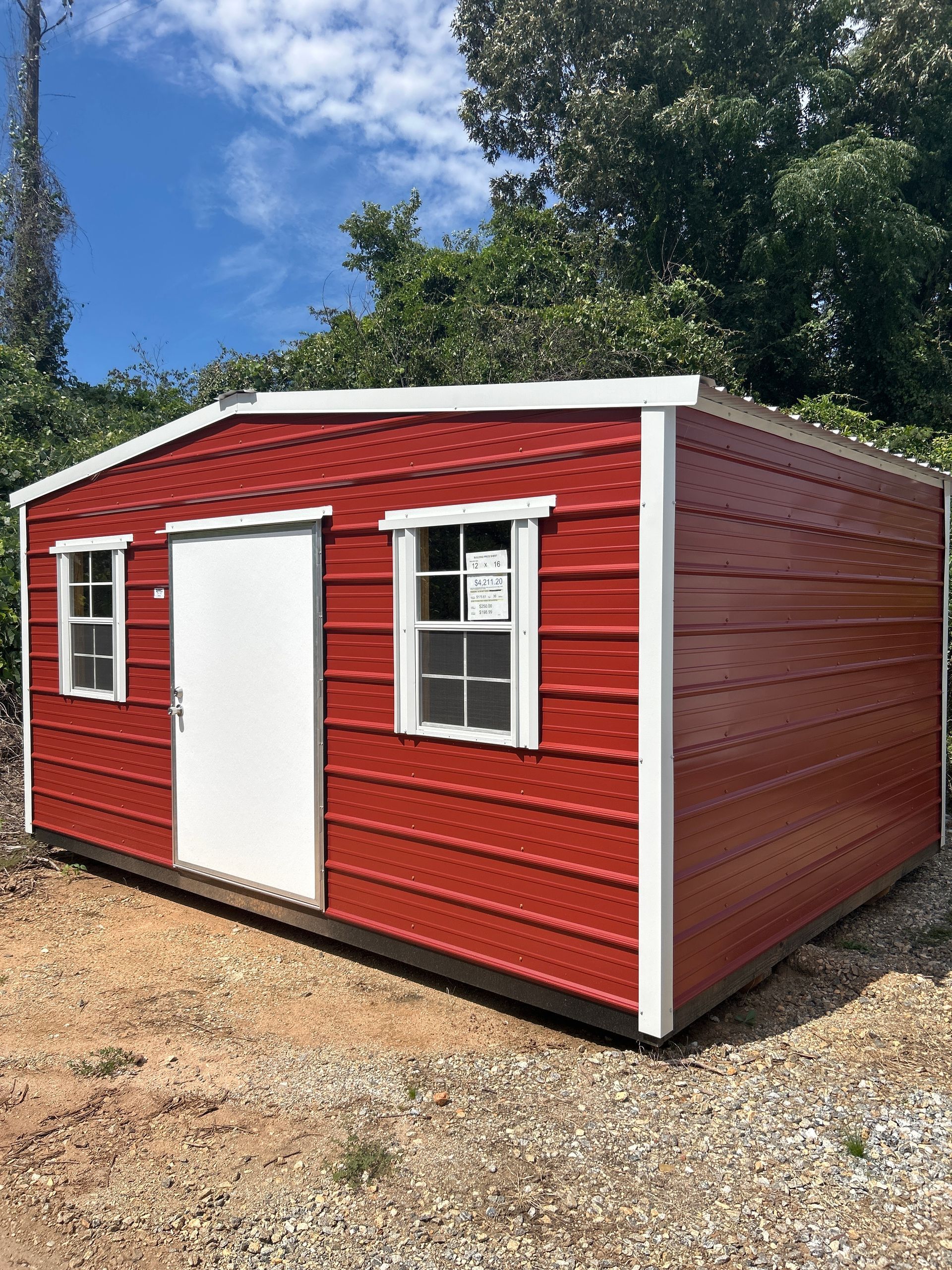 Red metal shed with white trim, door, and two windows, set against a backdrop of green foliage and a blue sky.