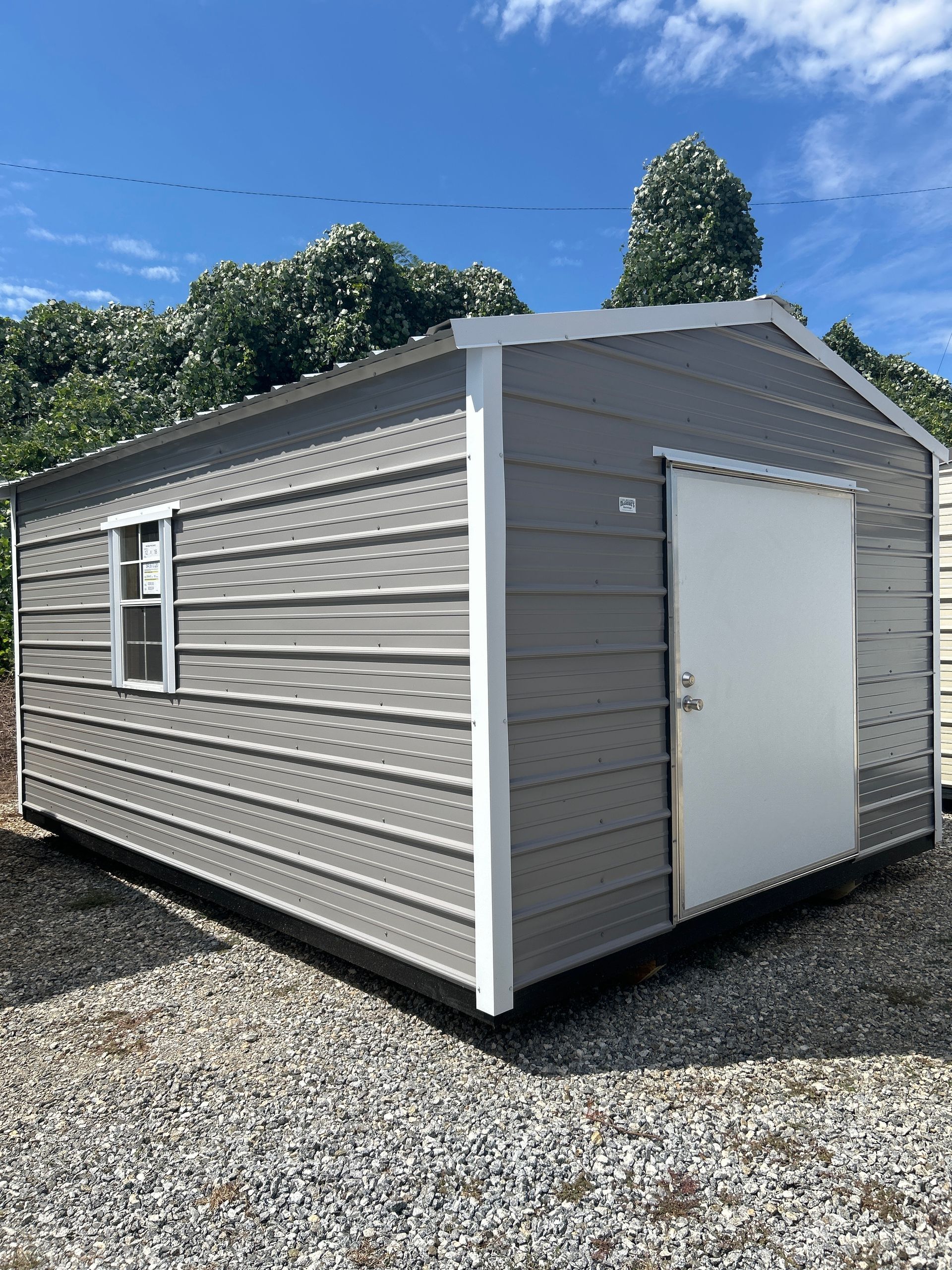 Gray metal storage shed with a white door and window, set on gravel.