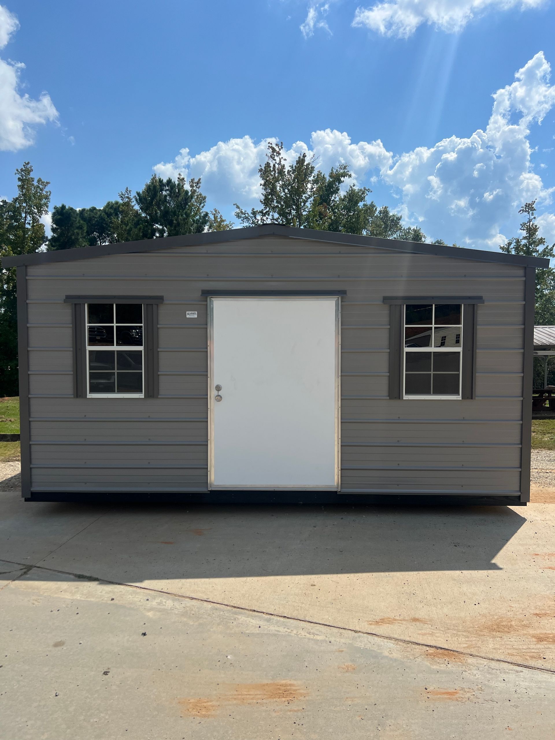 Gray shed with white door and windows against a blue sky with clouds.