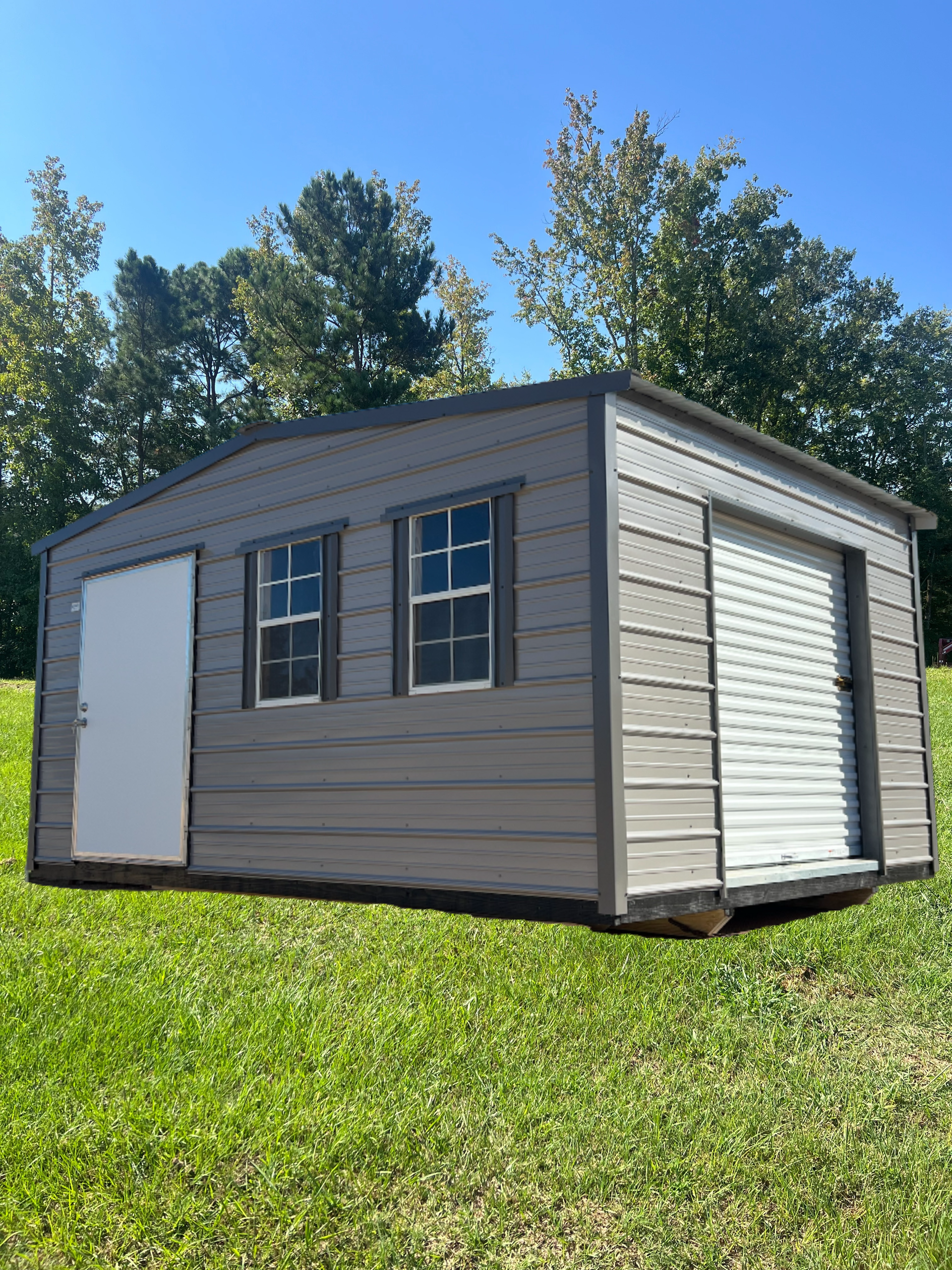 Gray shed with white door, windows, and garage door on green grass under blue sky.