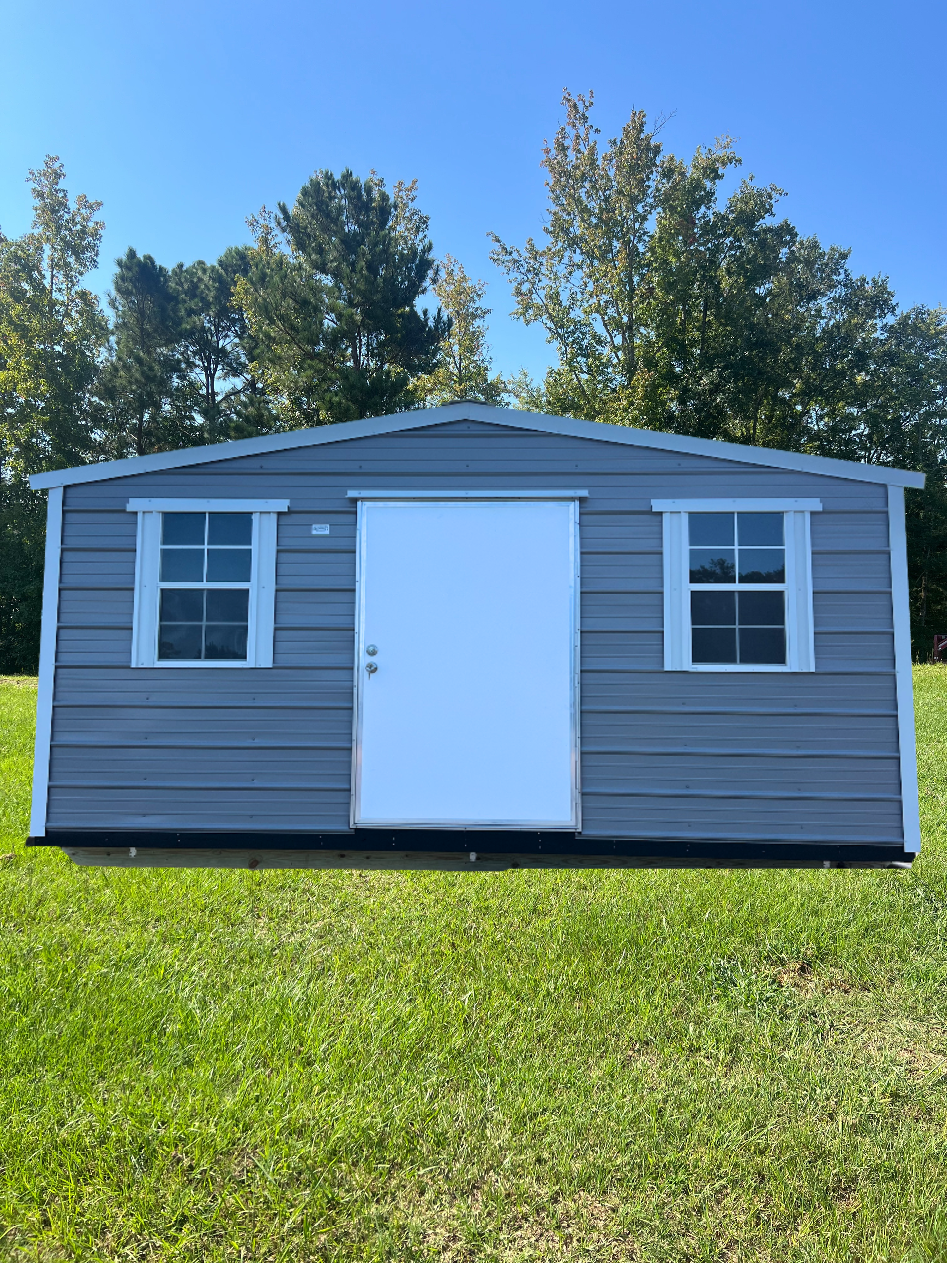 Gray shed with white door and window frames on a grassy lawn with trees in the background.