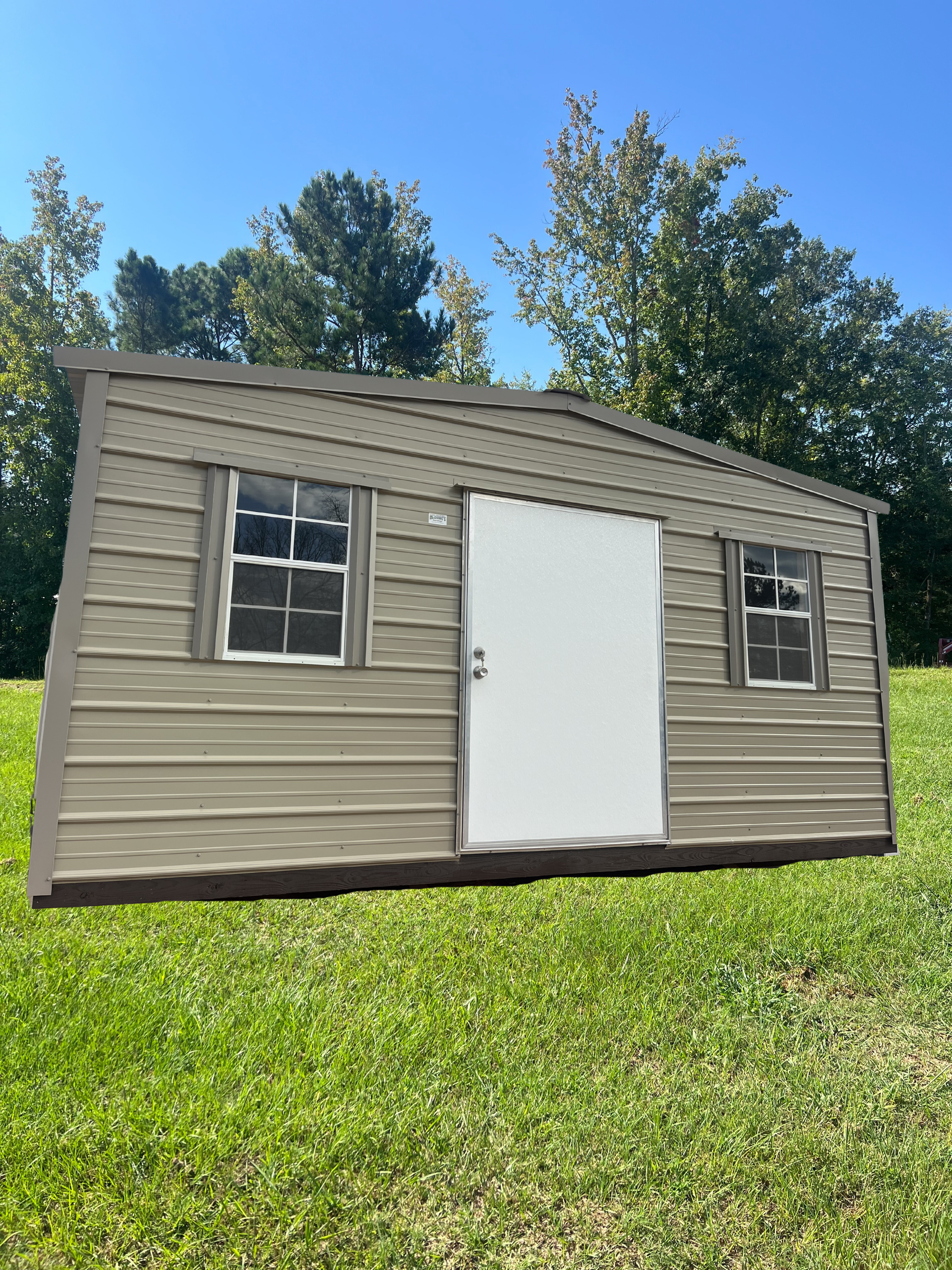 Tan shed with white door and windows on a grassy lawn with trees in the background.