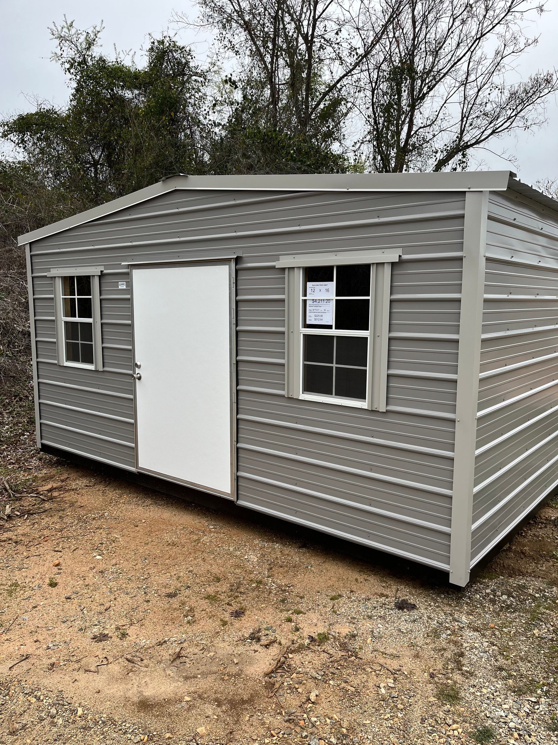 Gray metal shed with a white door and two windows, outdoors.