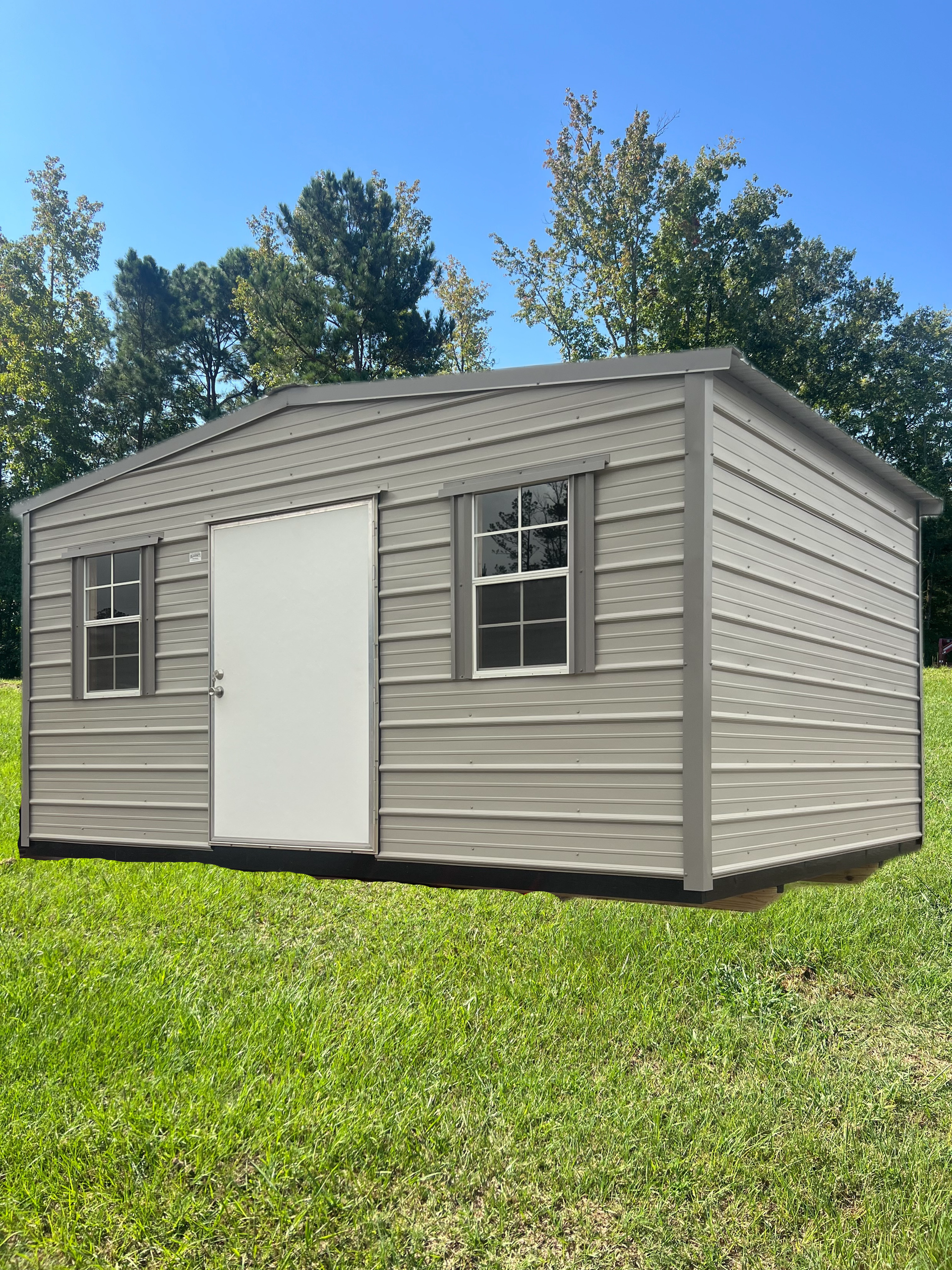 A gray storage shed with a white door and two windows sitting on a grassy lawn under a clear blue sky.