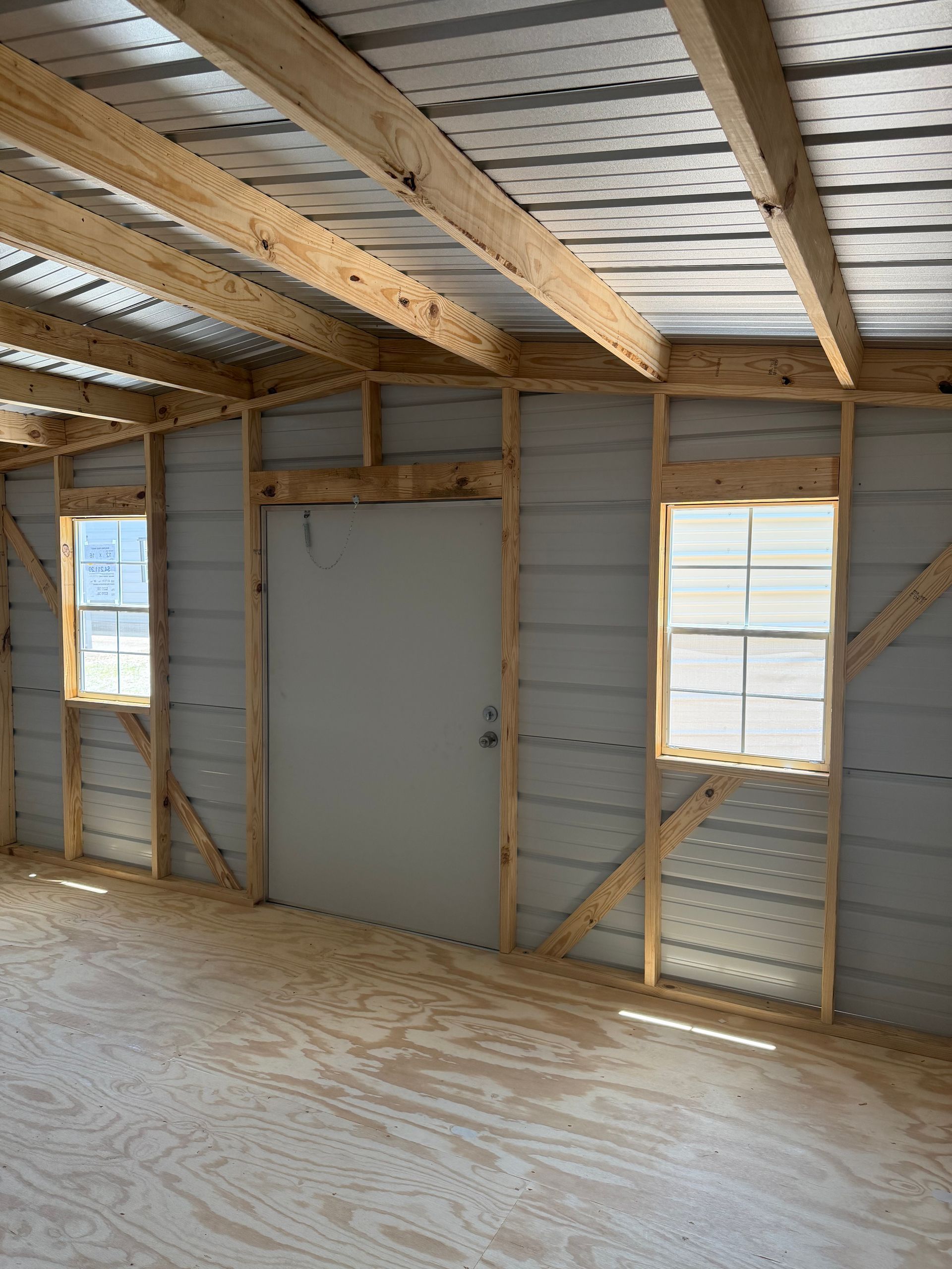 Unfinished room with exposed studs, plywood floor, and two windows on the right wall.