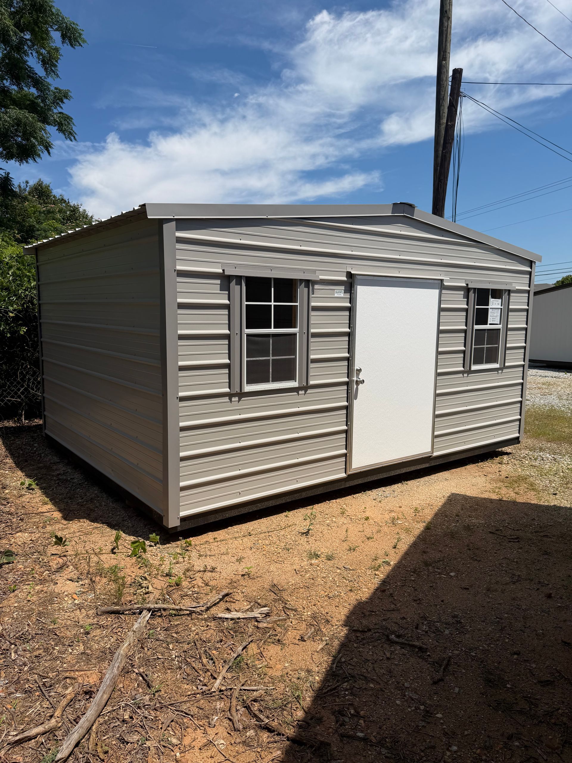 Small gray shed with a white door and windows, sitting on a gravel lot under a blue sky.