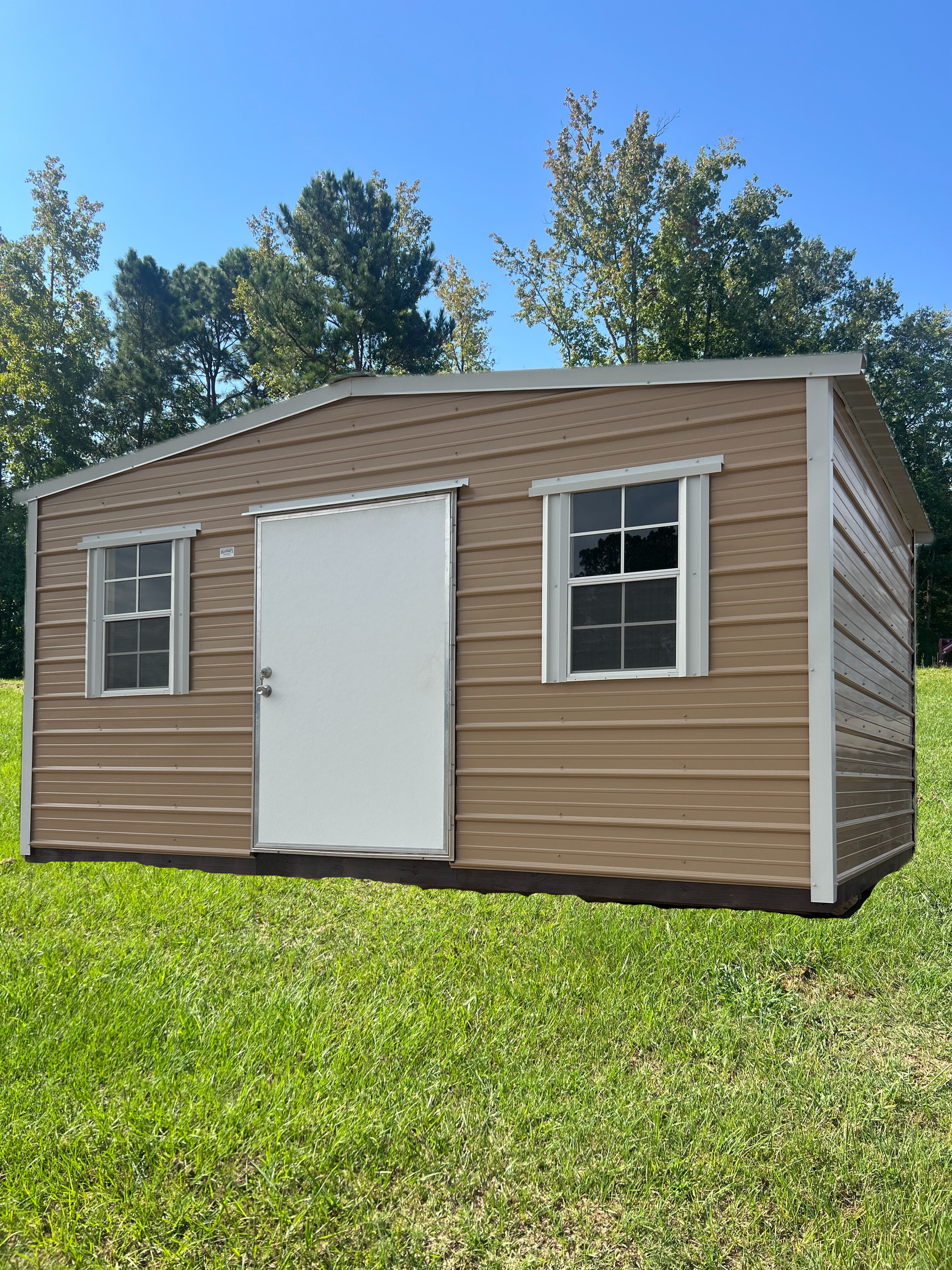 A tan, metal-sided storage shed with a white door and two windows sitting on green grass against a backdrop of trees.