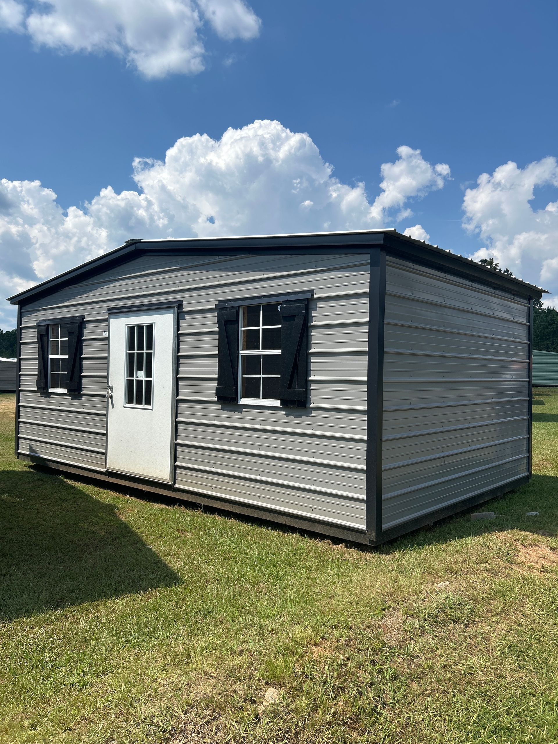 Gray shed with black trim, white door, black shutters, and a cloudy blue sky.