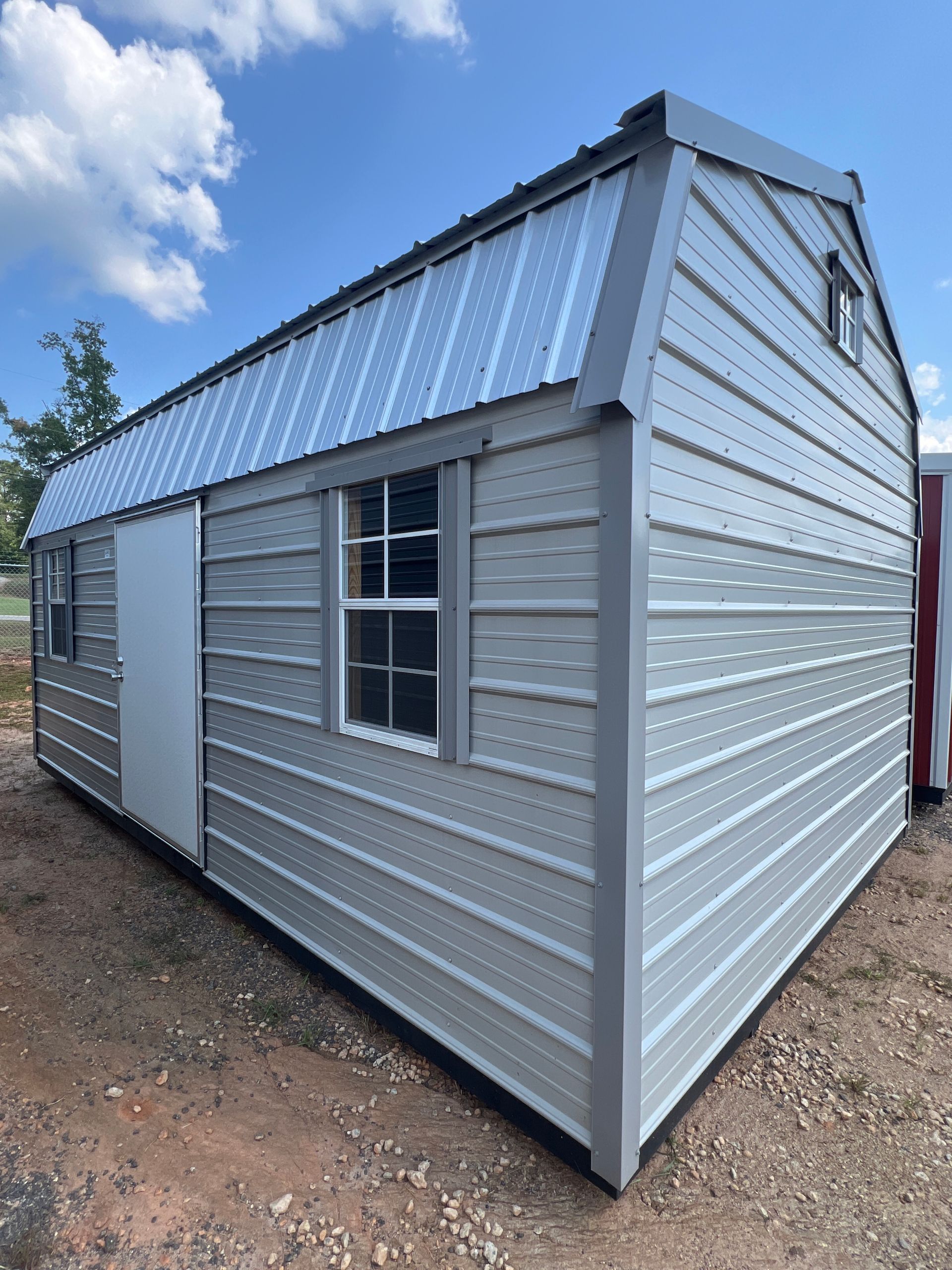Gray barn-style shed with metal roof, door, two windows, and triangular window in the gable, set outdoors.