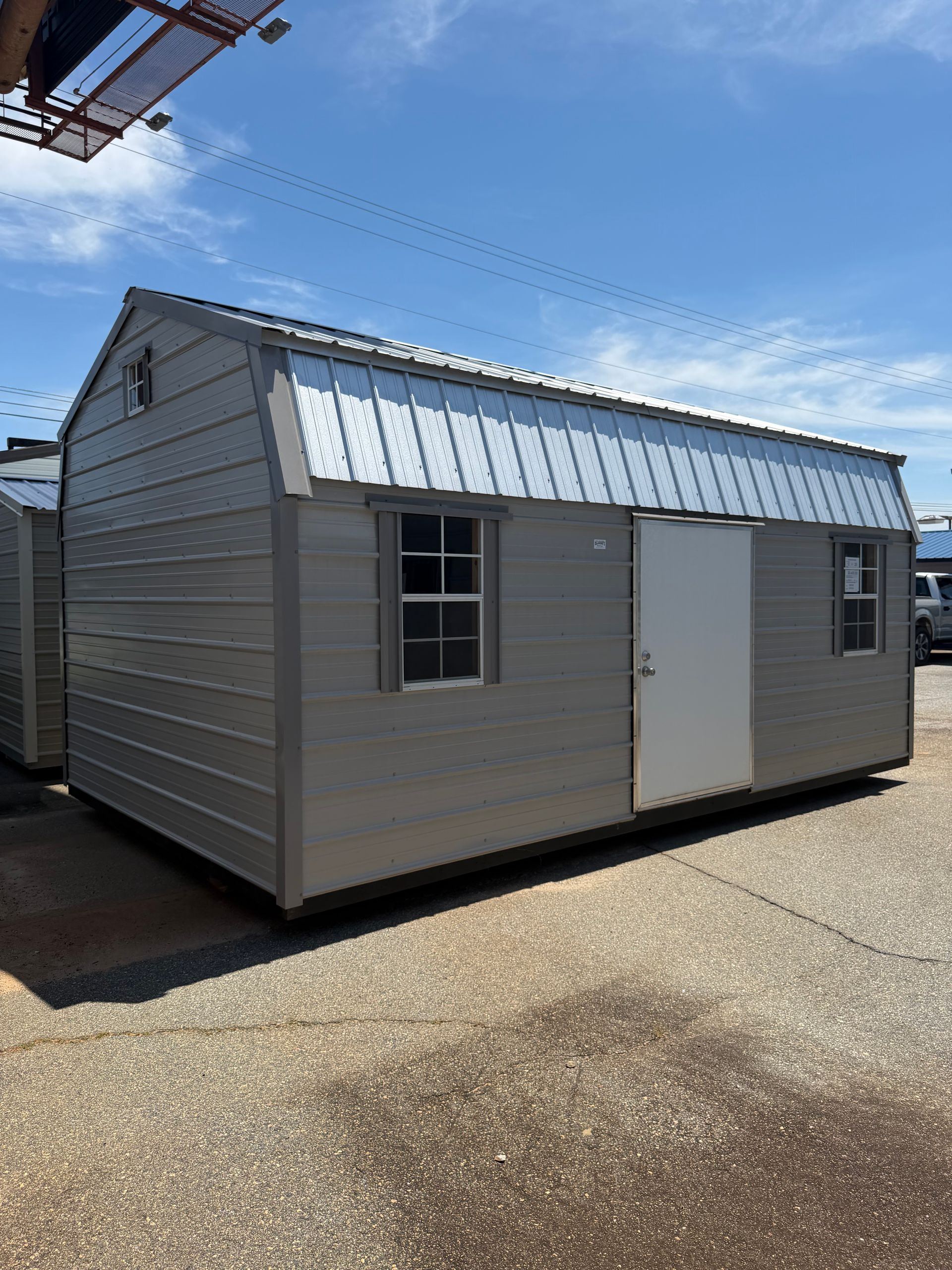 Small gray shed with a metal roof and white door on a gravel lot under a clear blue sky