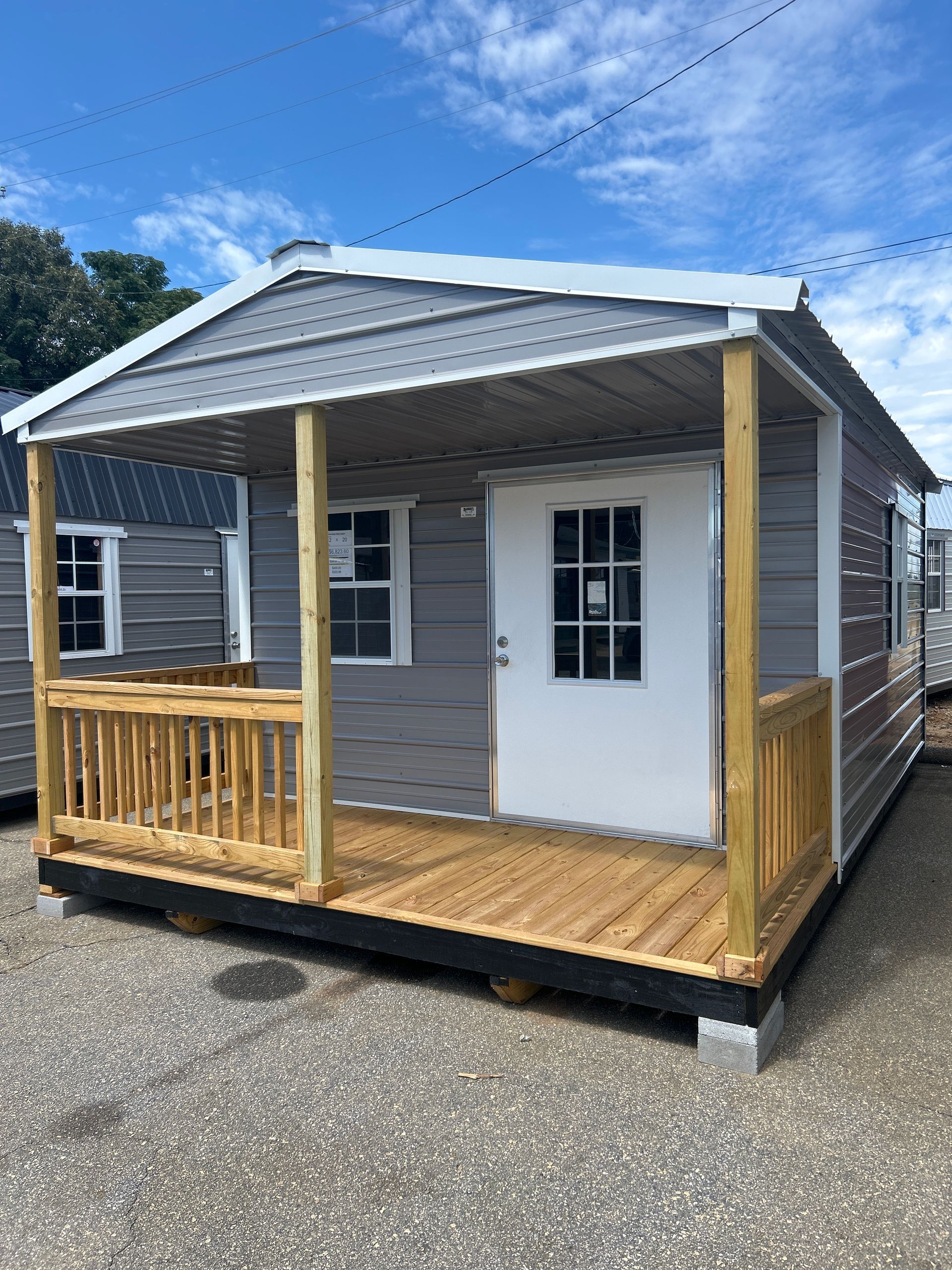Small gray shed with a porch and wooden railings, sitting on concrete blocks, against a blue sky.