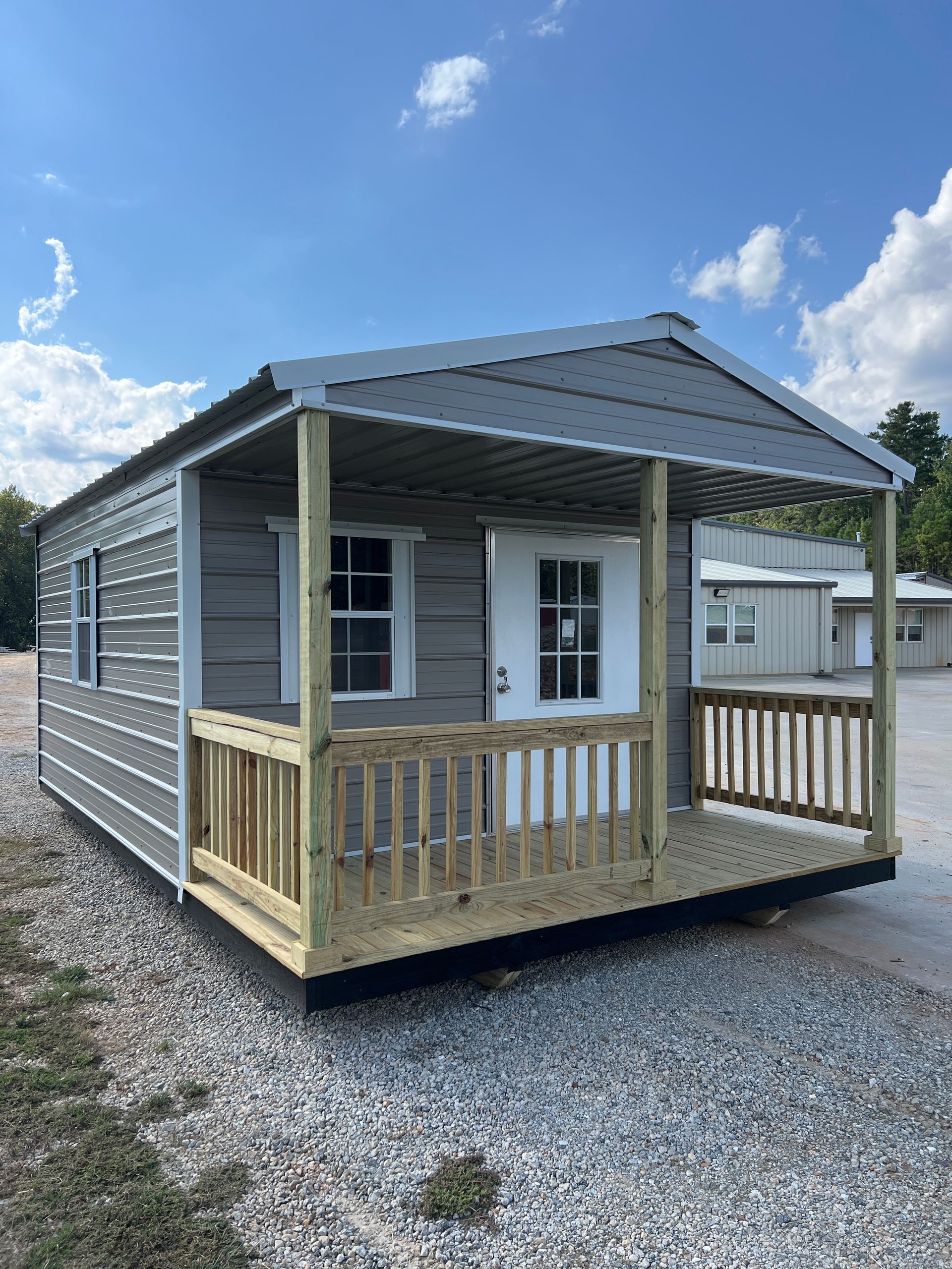 Gray tiny house with porch, white door, and small windows, on a gravel lot under a blue sky.
